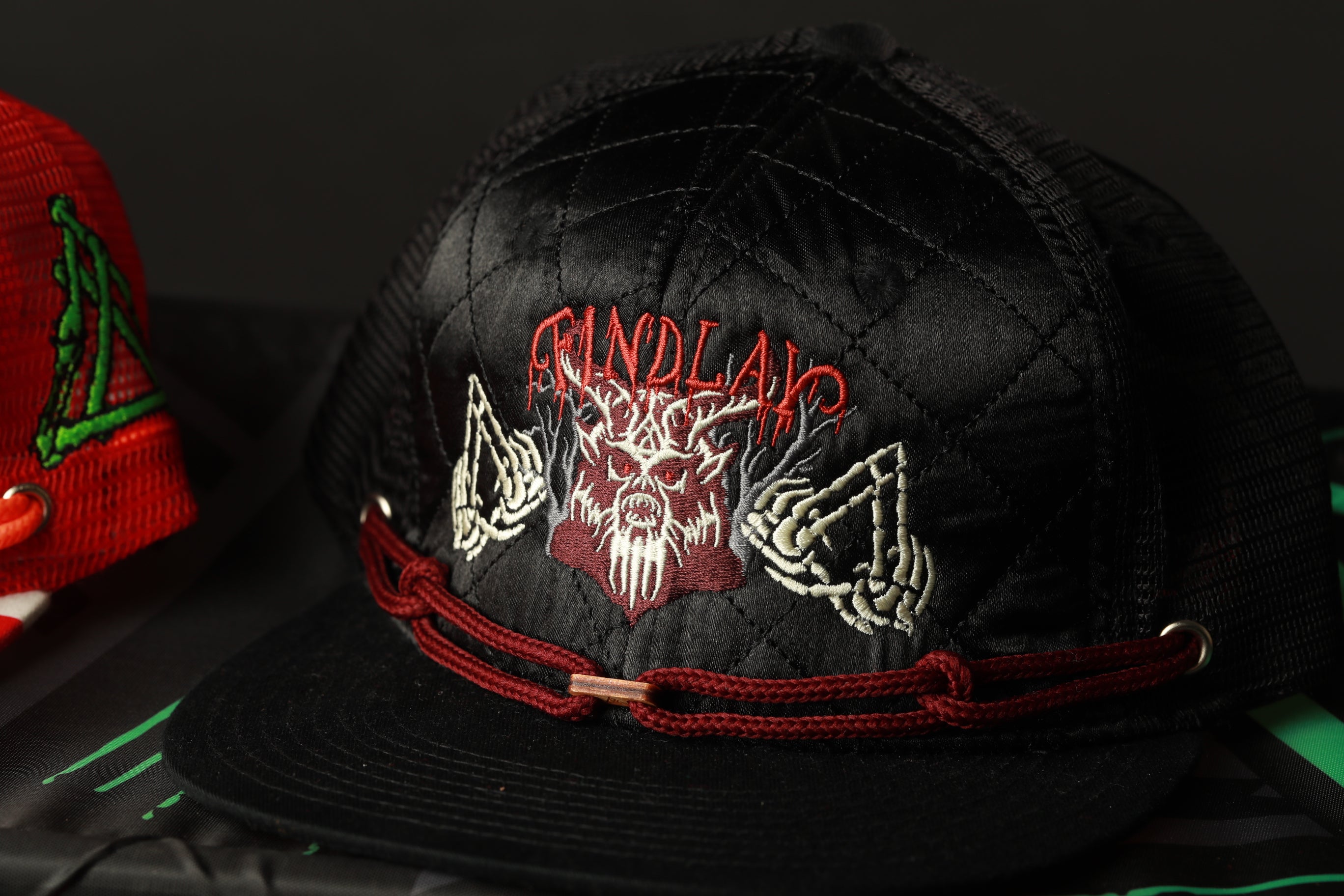 A black baseball cap with red and white embroidered designs, including a skull and crossbones motif, and the word "FINDLAC" printed on it.