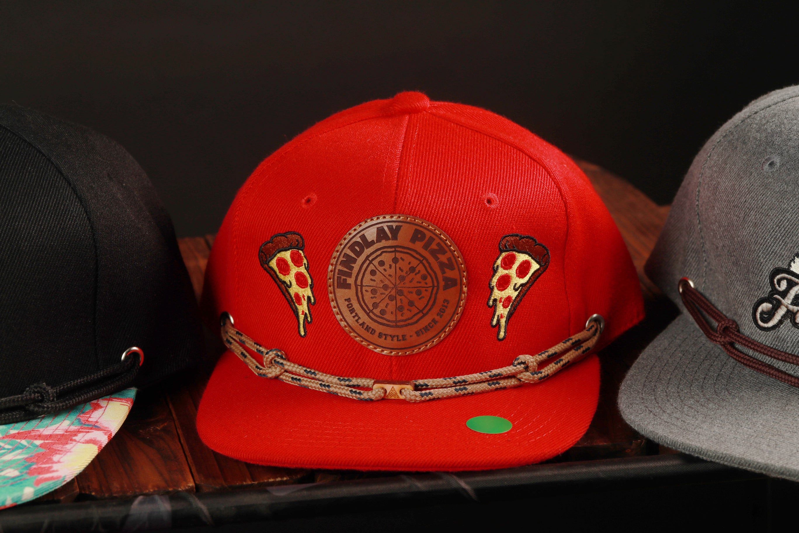 A red baseball cap with a pizza design, featuring two slices of pizza on either side of a circular logo, is displayed among other hats on a wooden surface.