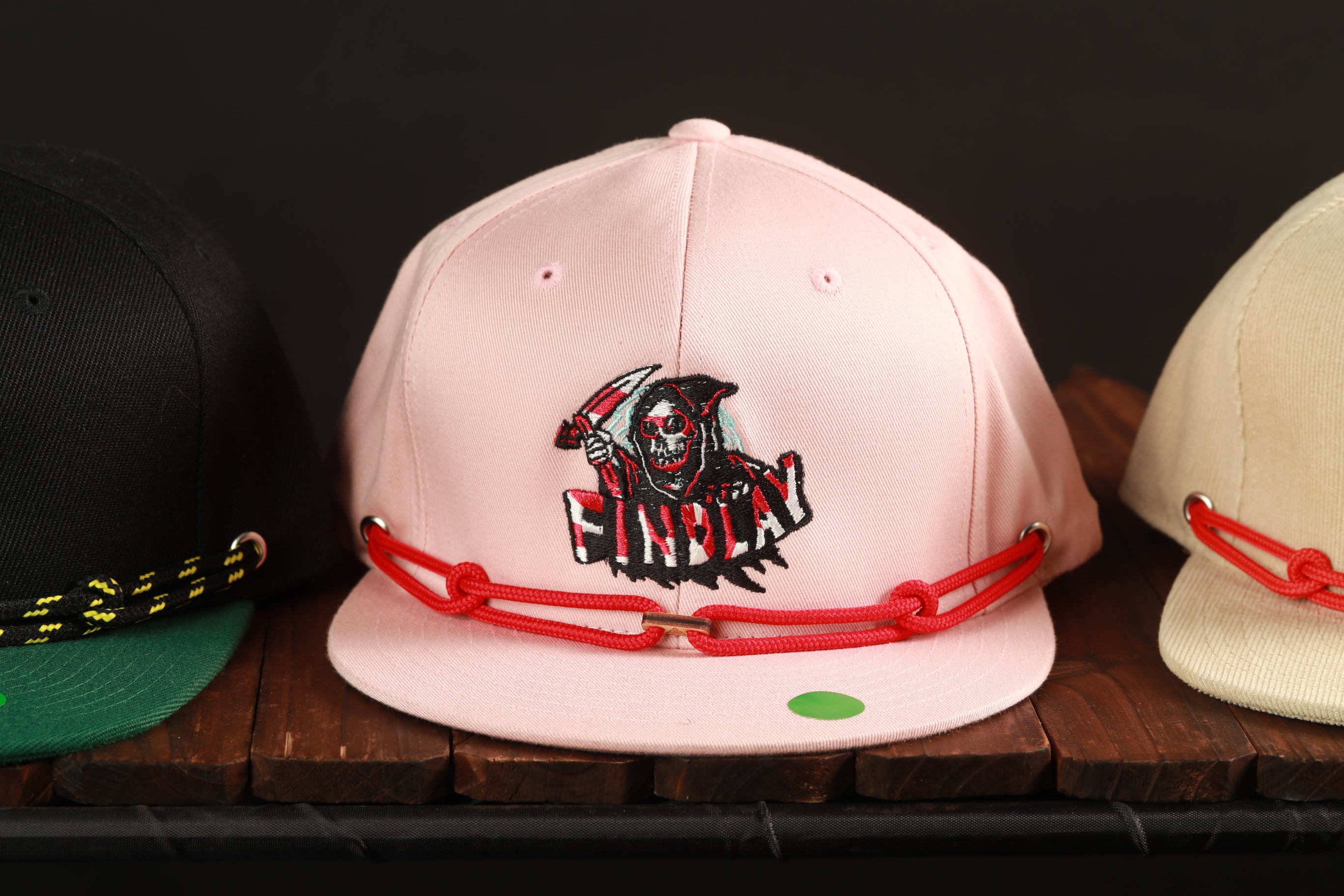 A pink baseball cap with a skull and crossbones design, a red string, and a green sticker on the front, displayed on a wooden surface.