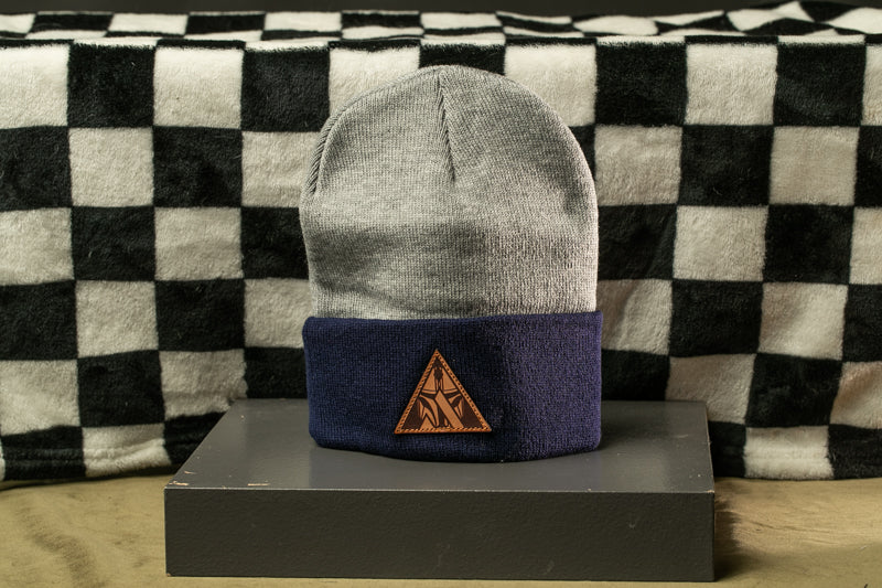 A gray and navy blue beanie with a brown triangle logo on the front, placed on a gray surface against a black and white checkered background.