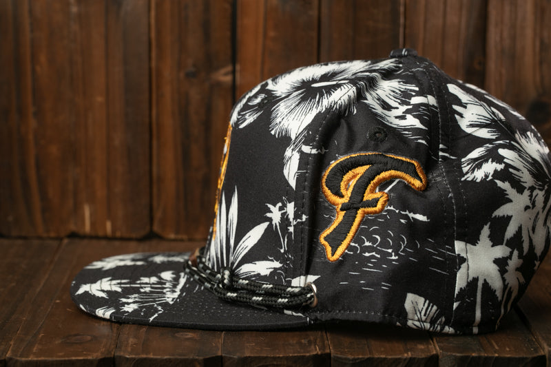 A black and white baseball cap with a floral pattern and a yellow "F" logo on the side, sitting on a wooden surface.