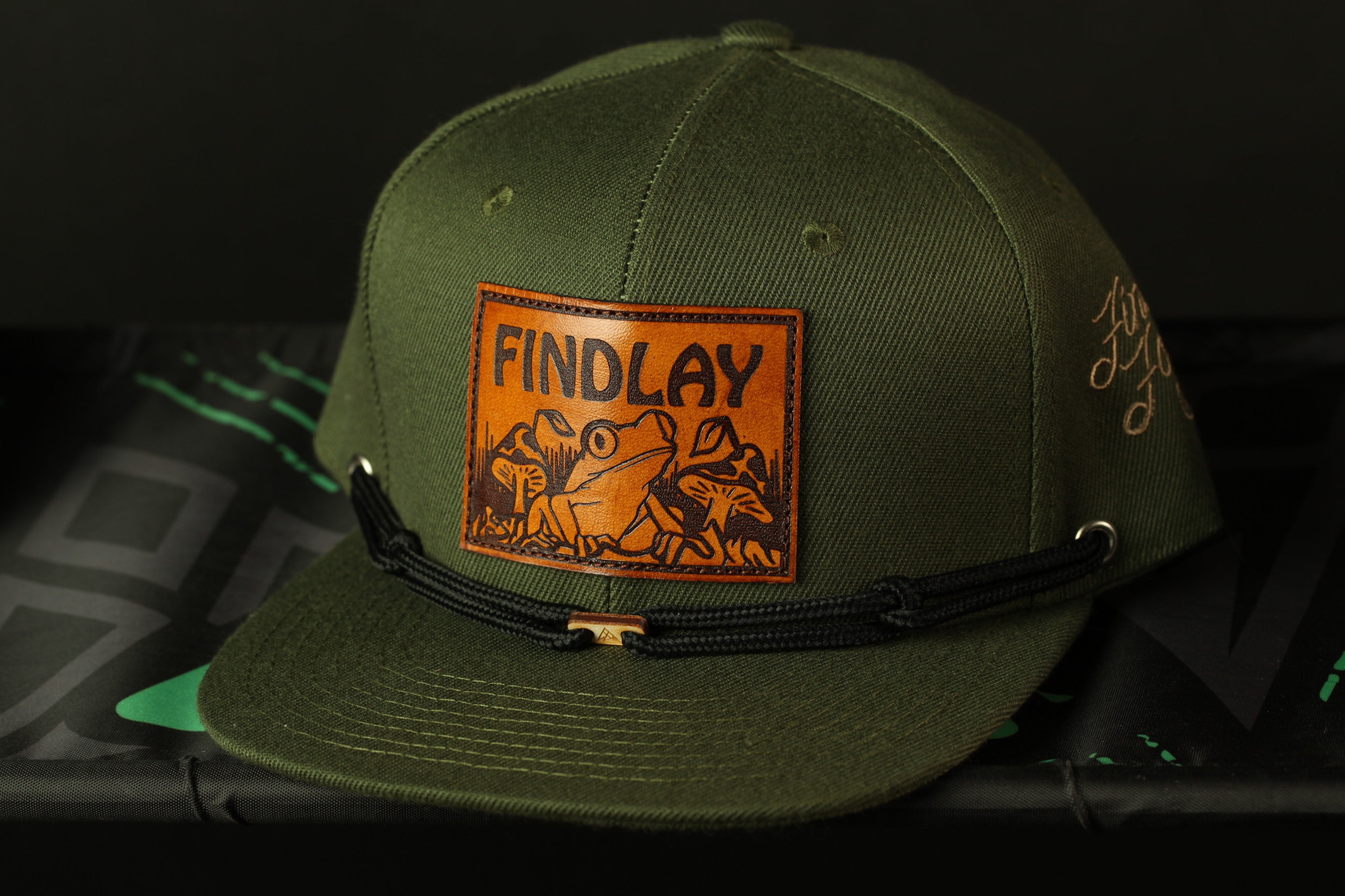 A green baseball cap with a leather patch featuring a frog and the word "FINDLAY" printed on it.