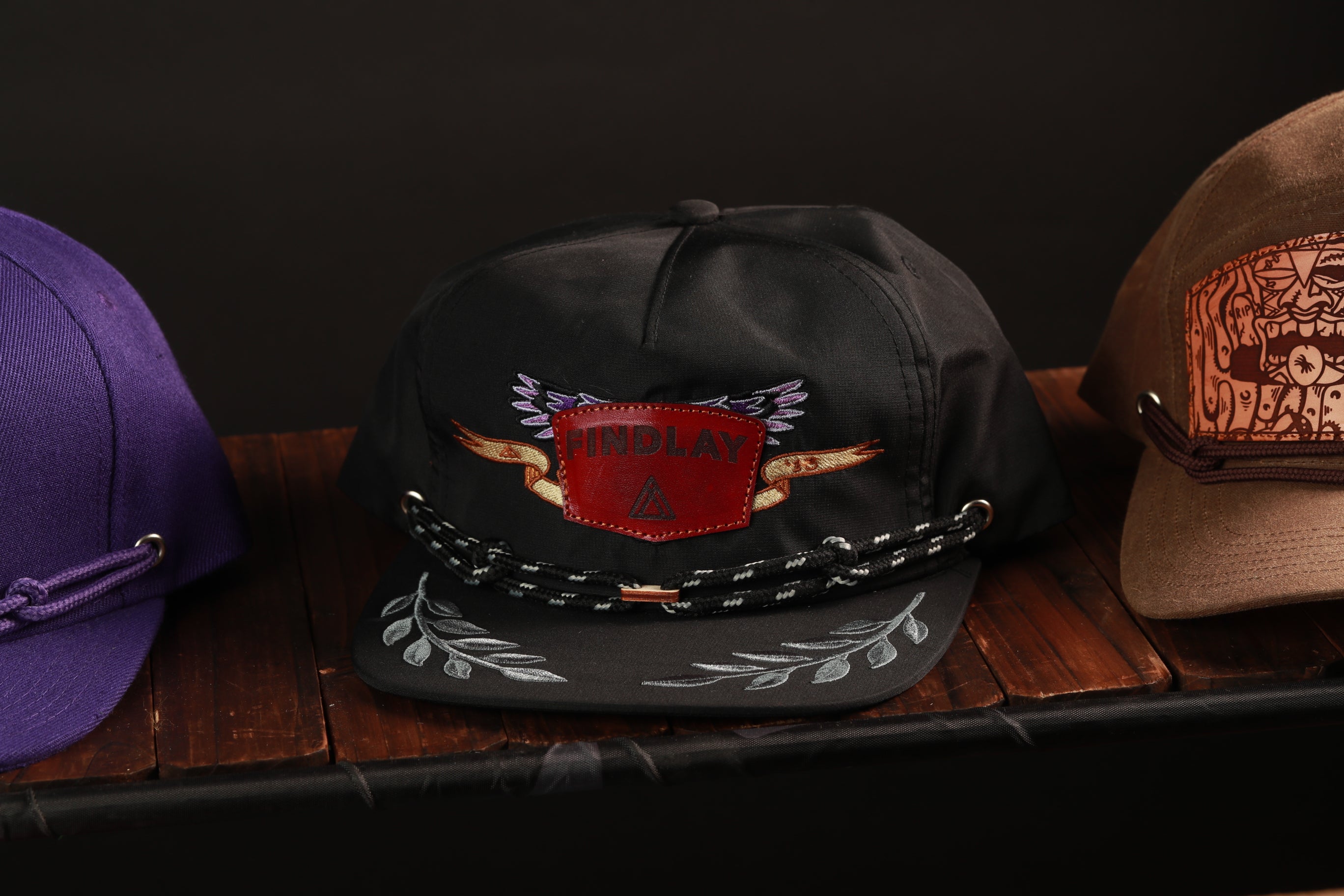 A collection of three baseball caps on a wooden surface, with one black cap featuring a red and white design and the word "INDAY" on it.