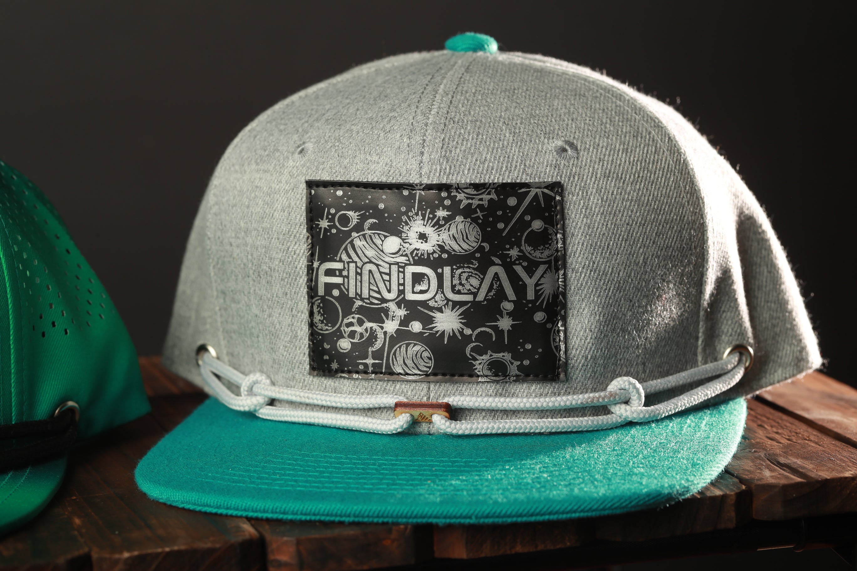 A gray baseball cap with a teal brim and a black patch on the front displaying the text "FIND LAY".