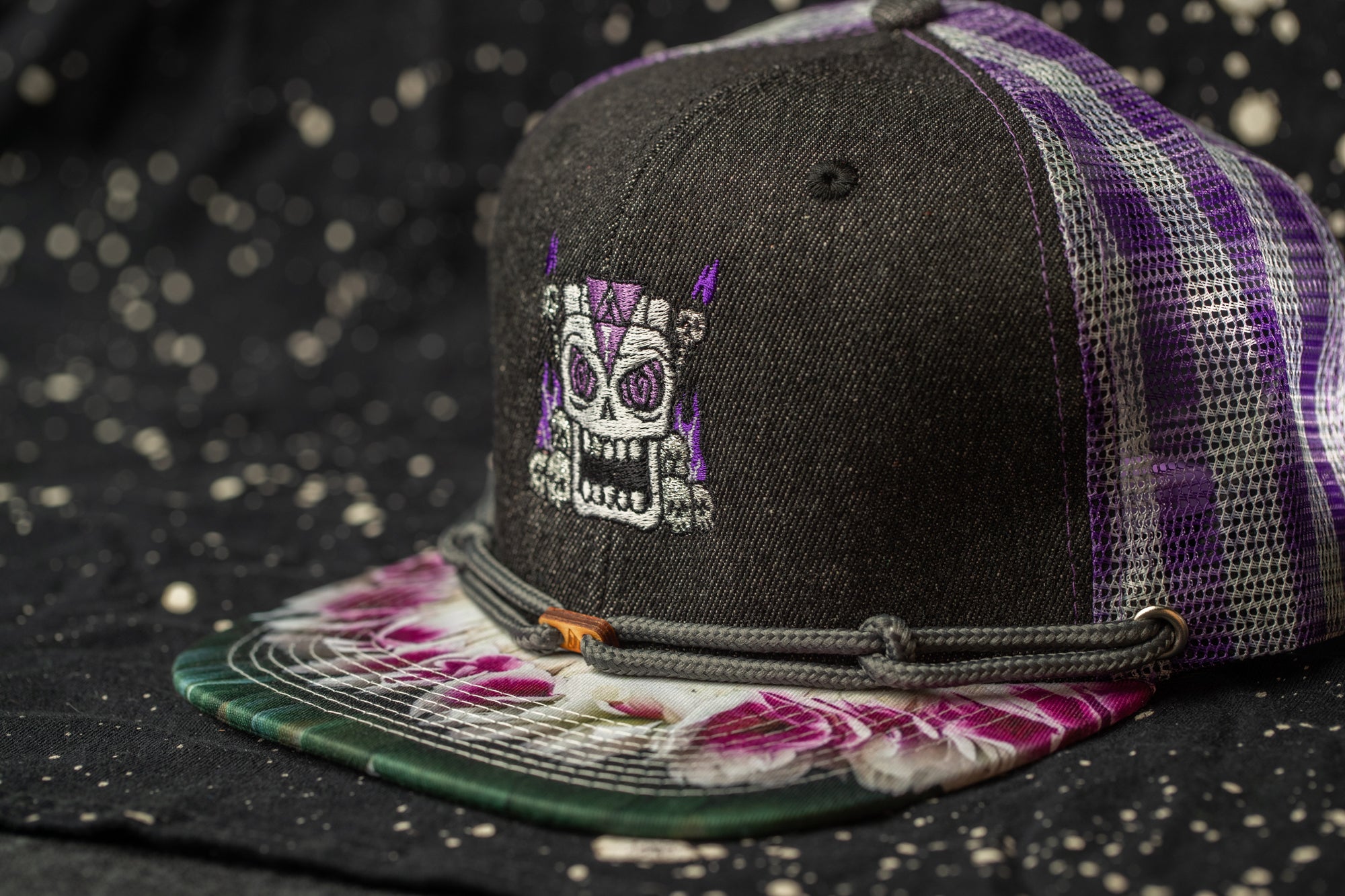 A black baseball cap with a purple and white patterned design and a skull embroidered on the front.