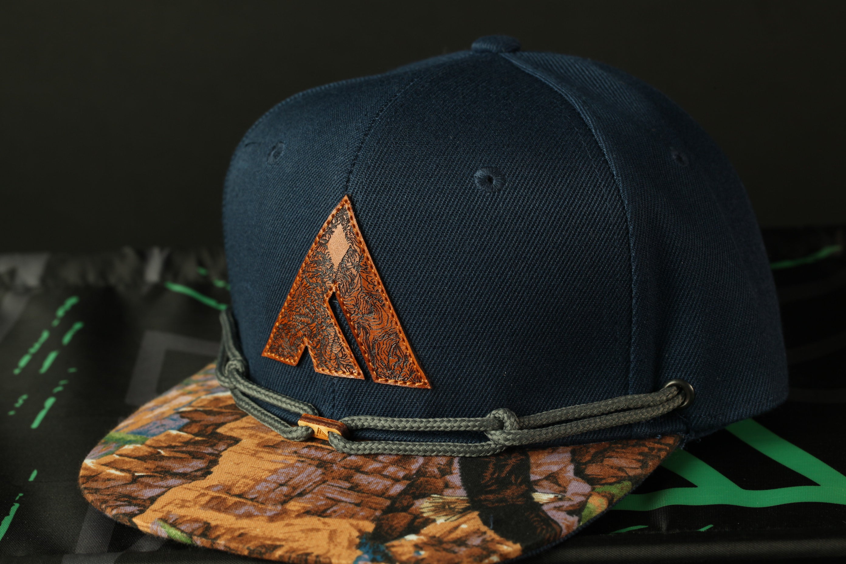 A navy blue baseball cap with a brown leather logo on the front, placed on a black surface with a green and black patterned fabric underneath.