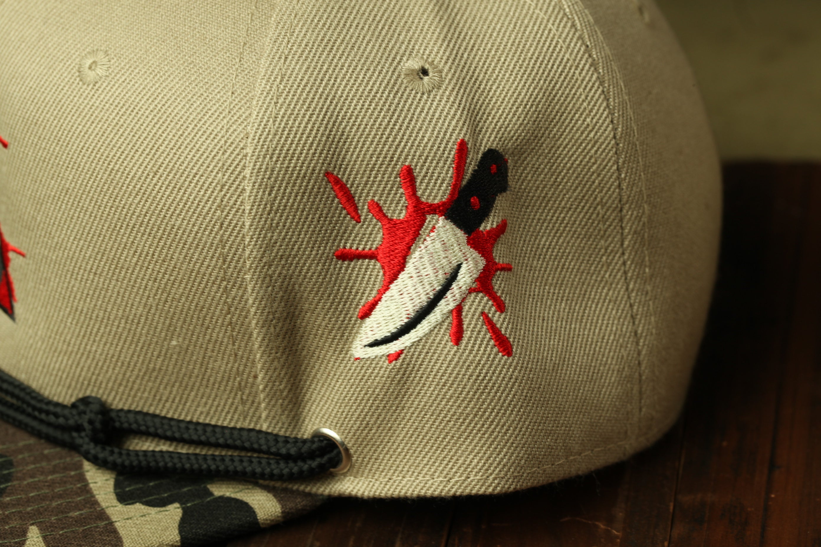 A close-up view of a beige baseball cap with a red and black embroidered design depicting a knife with a red splash of blood.