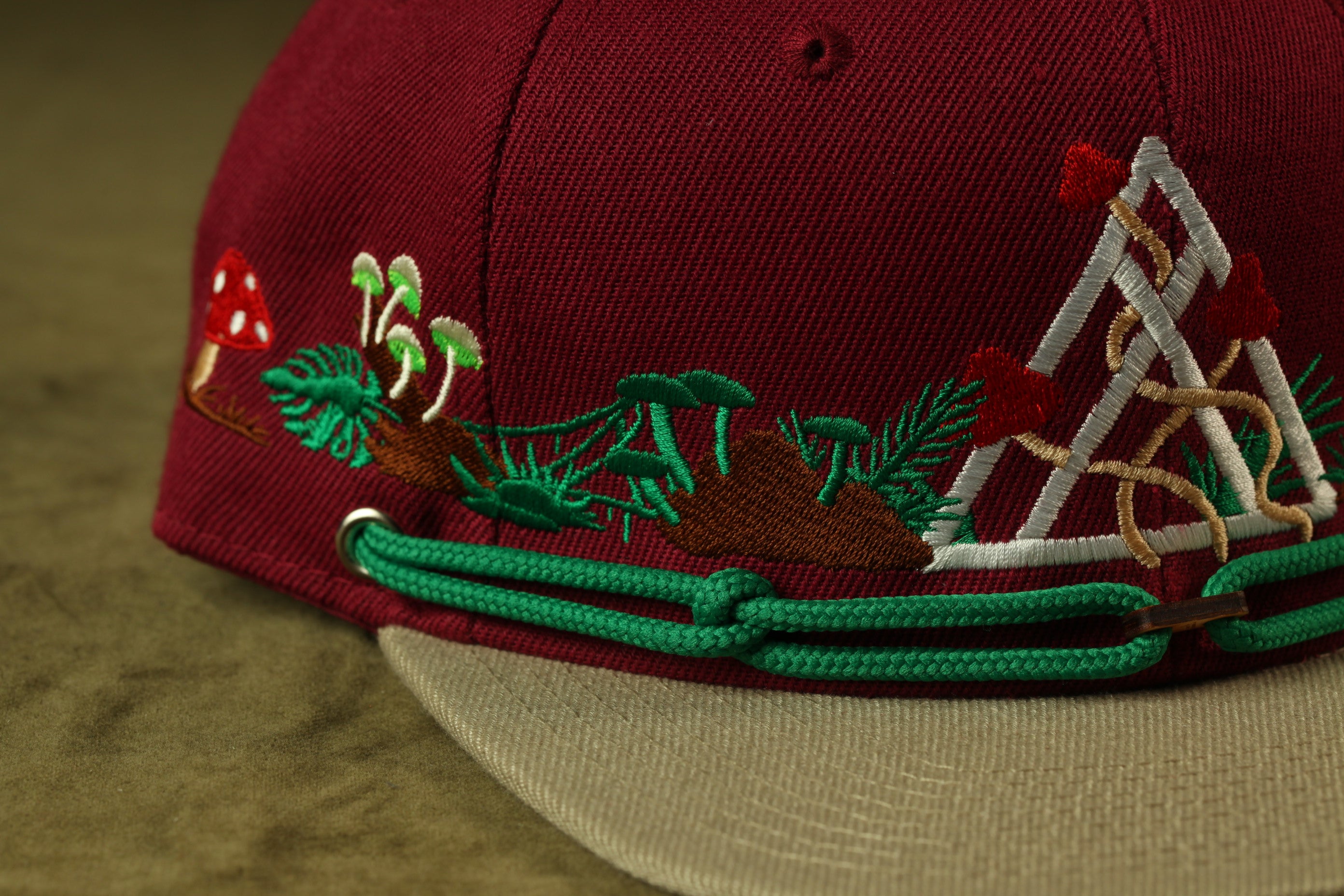 A close-up view of a burgundy baseball cap with intricate embroidered designs, including mushrooms, leaves, and geometric shapes.