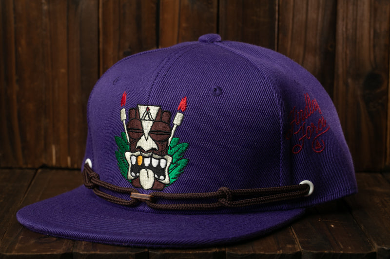 A purple baseball cap with a graphic design on the front, featuring a tiki mask and tropical foliage.