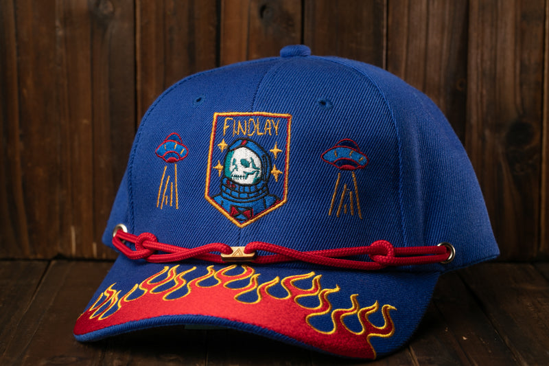 A blue baseball cap with a red and yellow flame design, featuring a skull and an alien motif, is displayed against a wooden background.