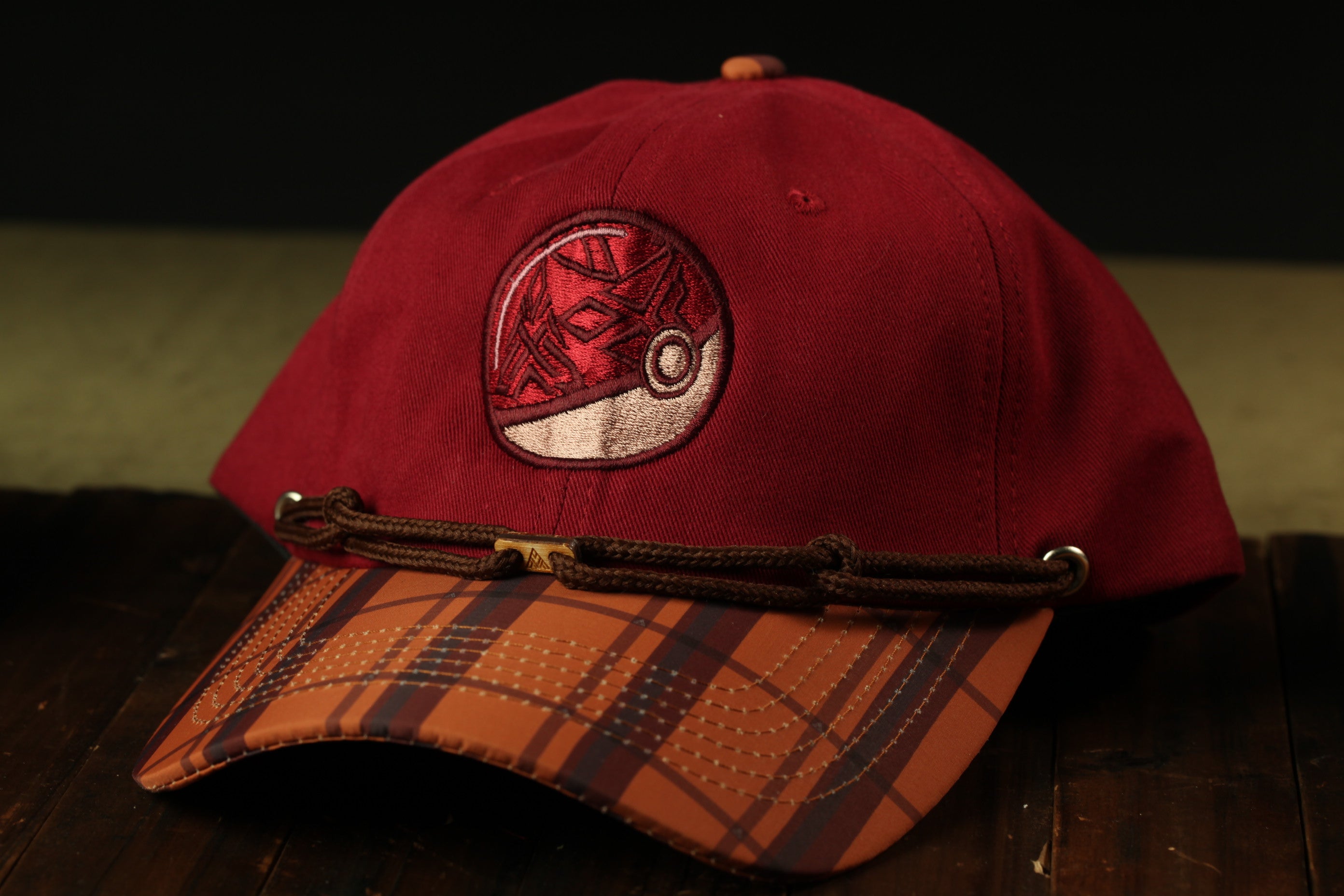 A red baseball cap with a plaid pattern and a Pokemon-themed logo is displayed on a wooden surface.