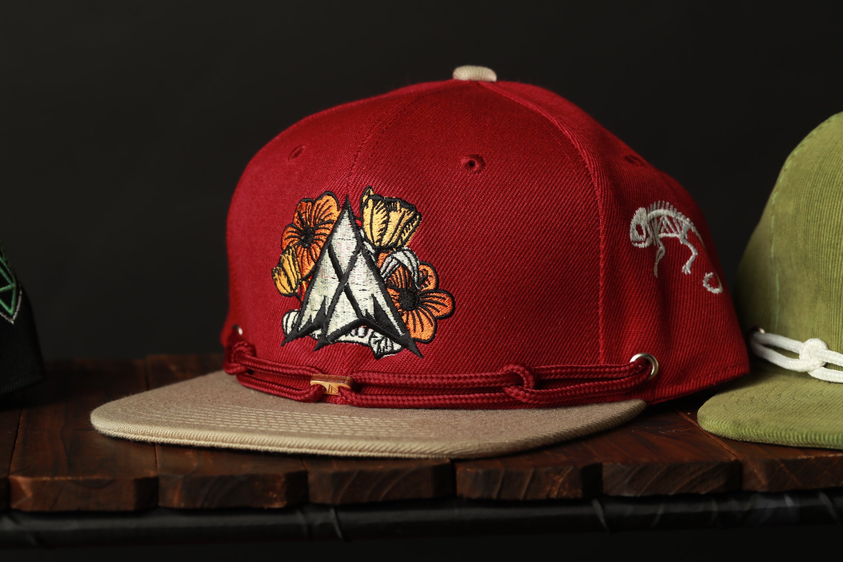 A red baseball cap with a floral design is displayed on a wooden surface, with another green baseball cap visible in the background.