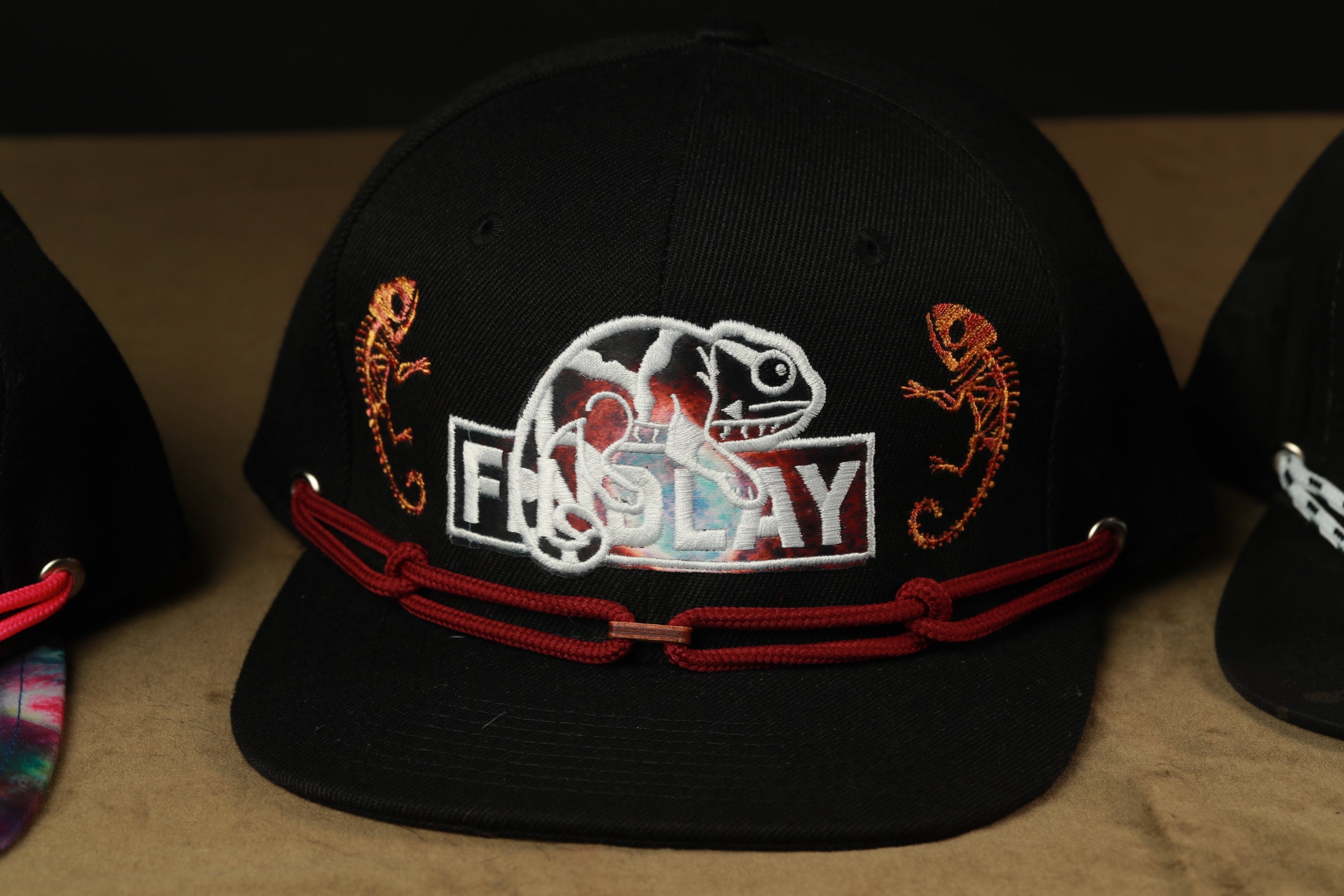 A black baseball cap with a red string and a white logo featuring a chameleon and the word "FIND PLAY".