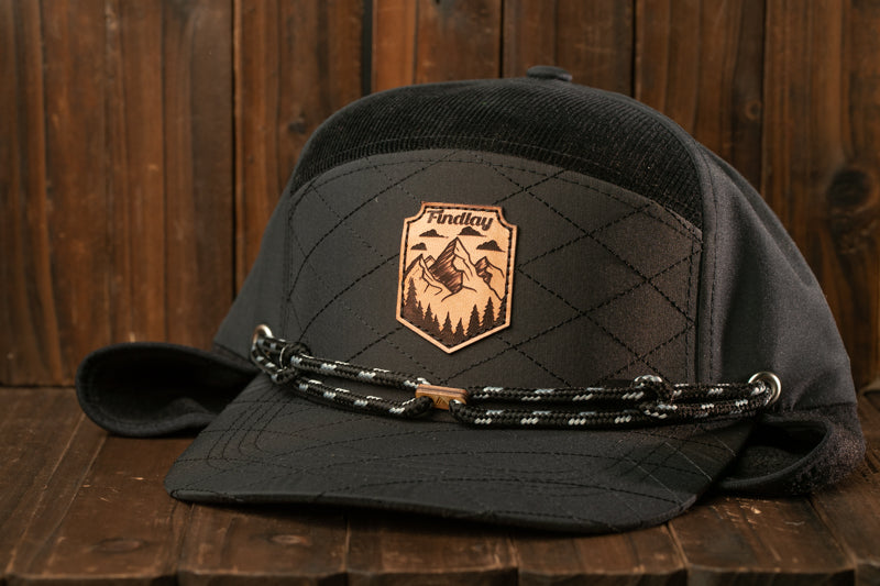 A black baseball cap with a mountain landscape patch and a black and white rope strap, placed on a wooden surface.