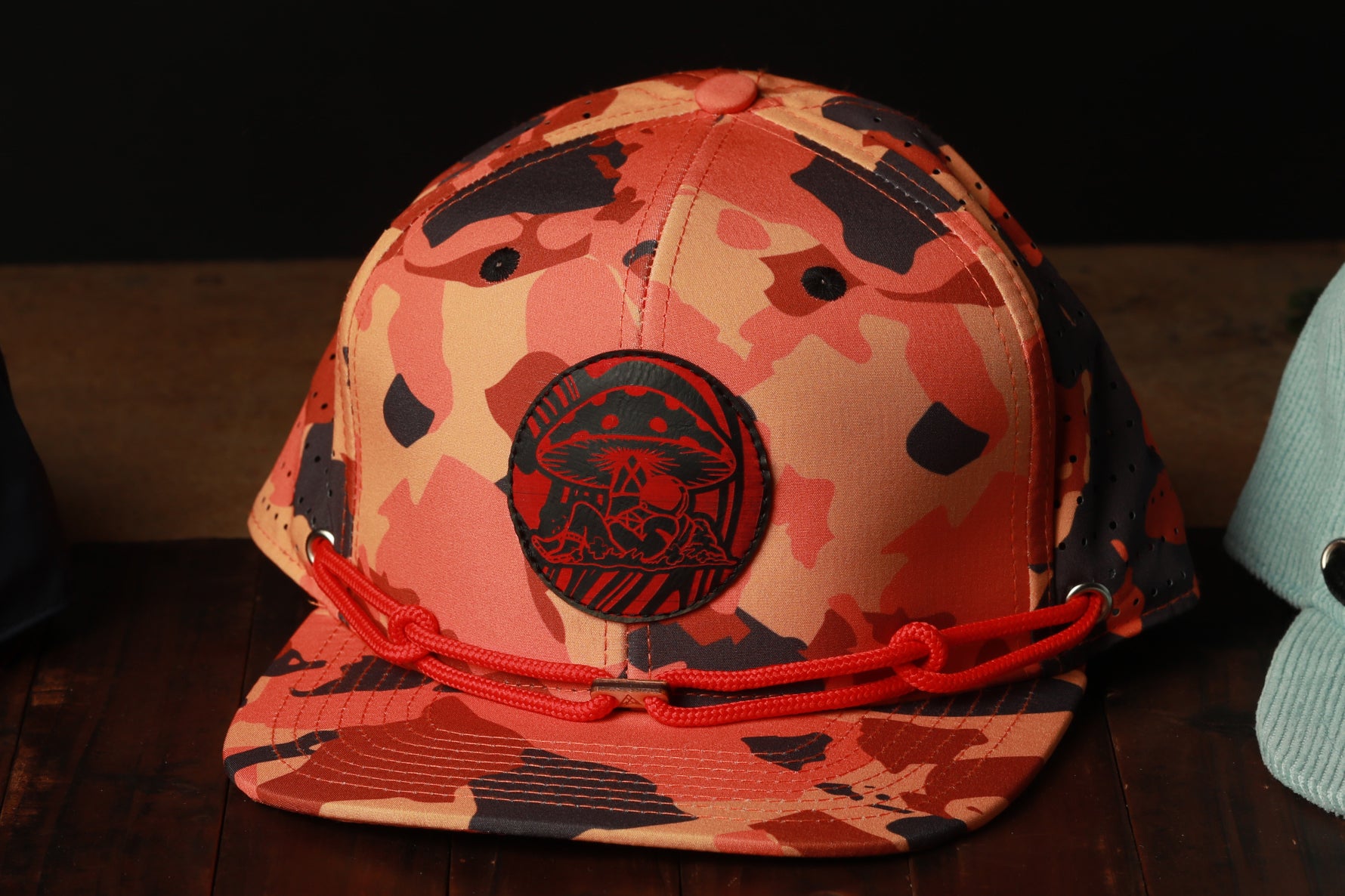 A camouflage baseball cap with a mushroom patch and a red chain strap is displayed on a wooden surface.