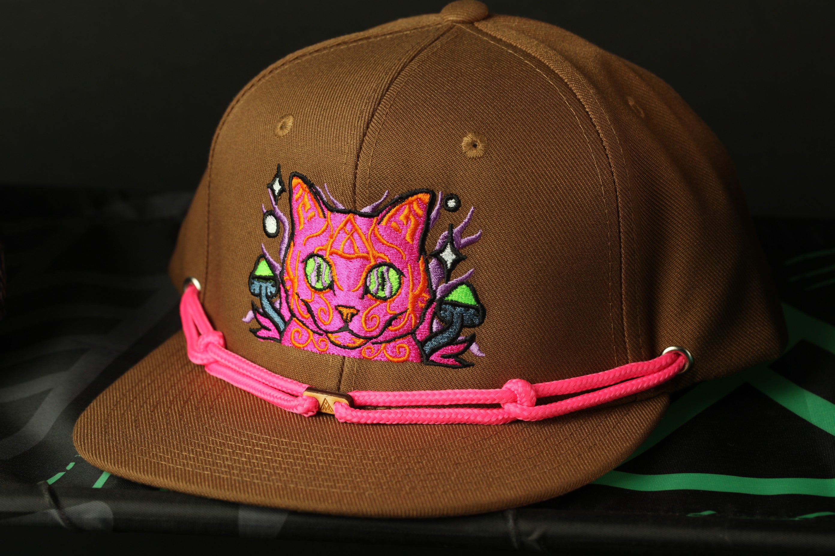 A brown baseball cap with a colorful cat embroidered on it, featuring a pink braided strap.