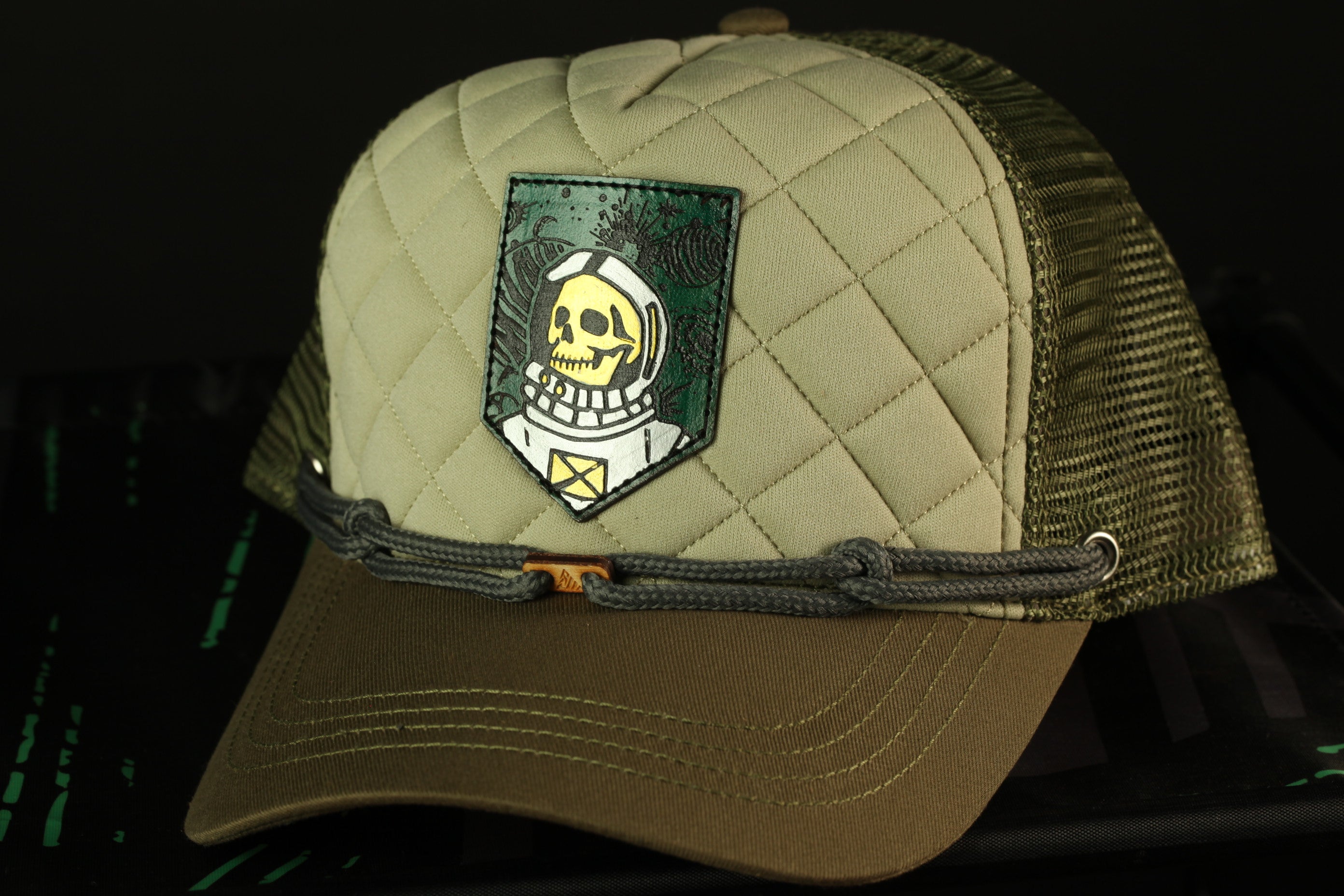 A beige baseball cap with a green and white skull patch on the front, featuring a rope strap.