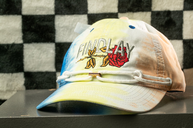 A tie-dye baseball cap with the word "FINDLAY" embroidered on it, sitting on a table against a checkered background.