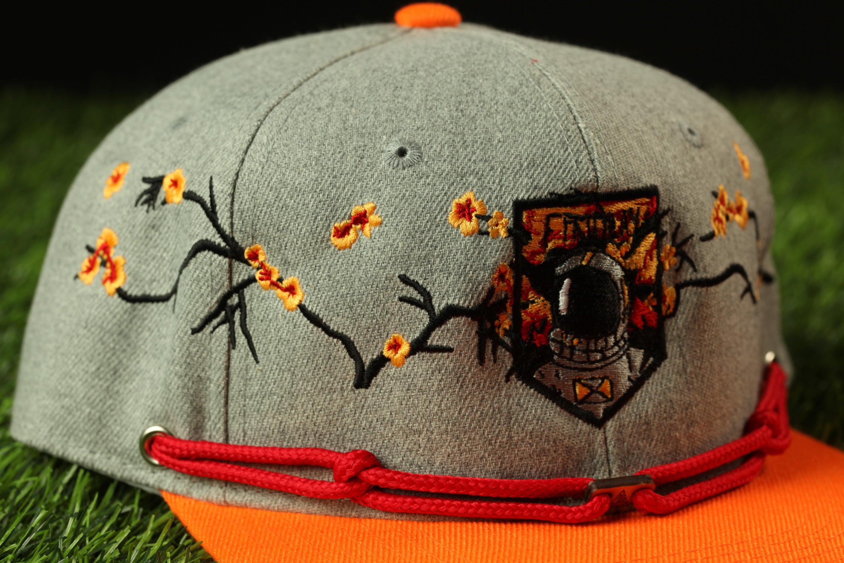 A gray baseball cap with a red string and a black and orange patch featuring a graphic design on the front.
