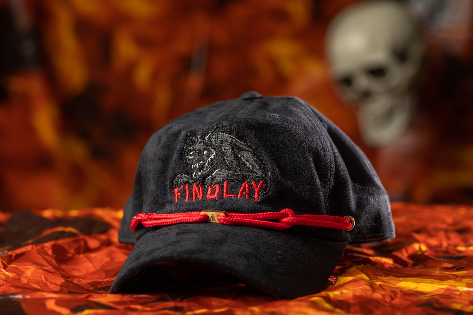 A black baseball cap with a red string and the word "FINDOLAY" printed on it, placed on a red and orange patterned surface.
