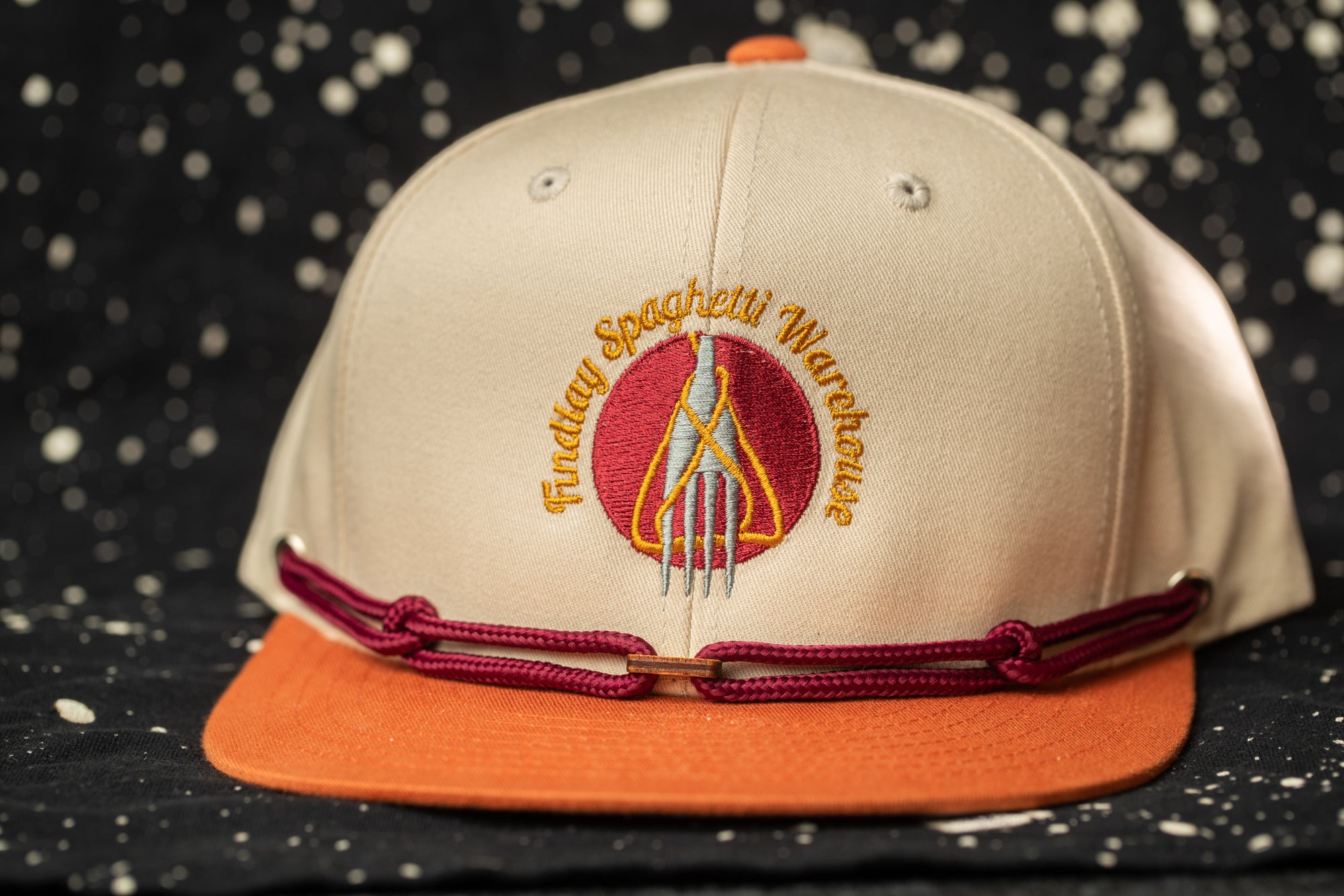 A beige baseball cap with a maroon band and a logo on the front, against a black background with white speckles.