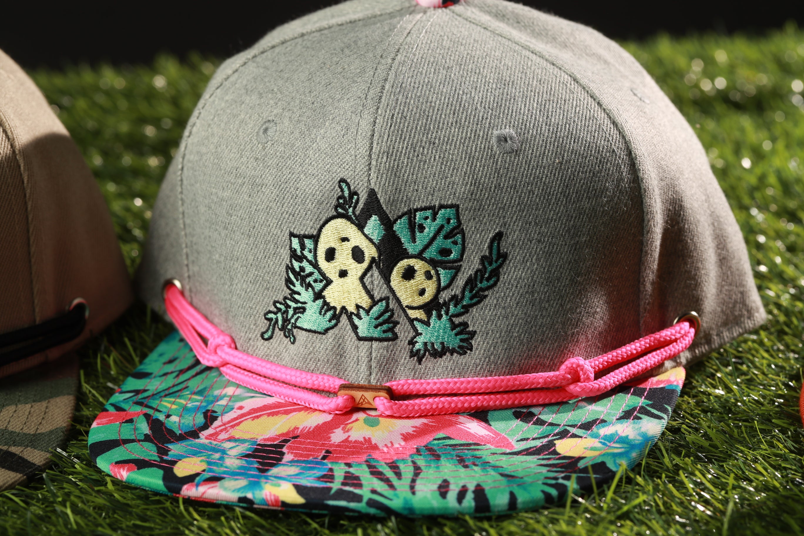 A gray baseball cap with a floral pattern and a pink braided strap, placed on a grassy surface.