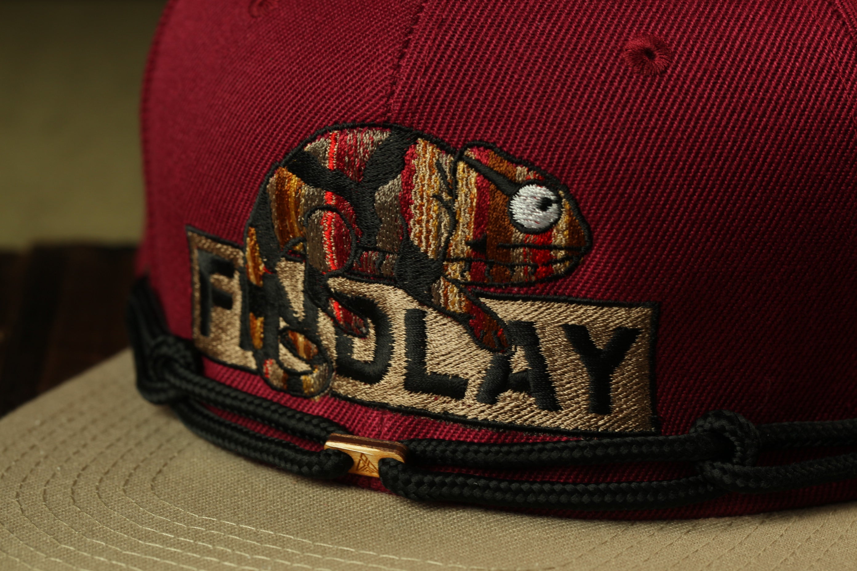 A close-up of a maroon baseball cap with a colorful, embroidered design featuring a chameleon and the word "FIND LAY".