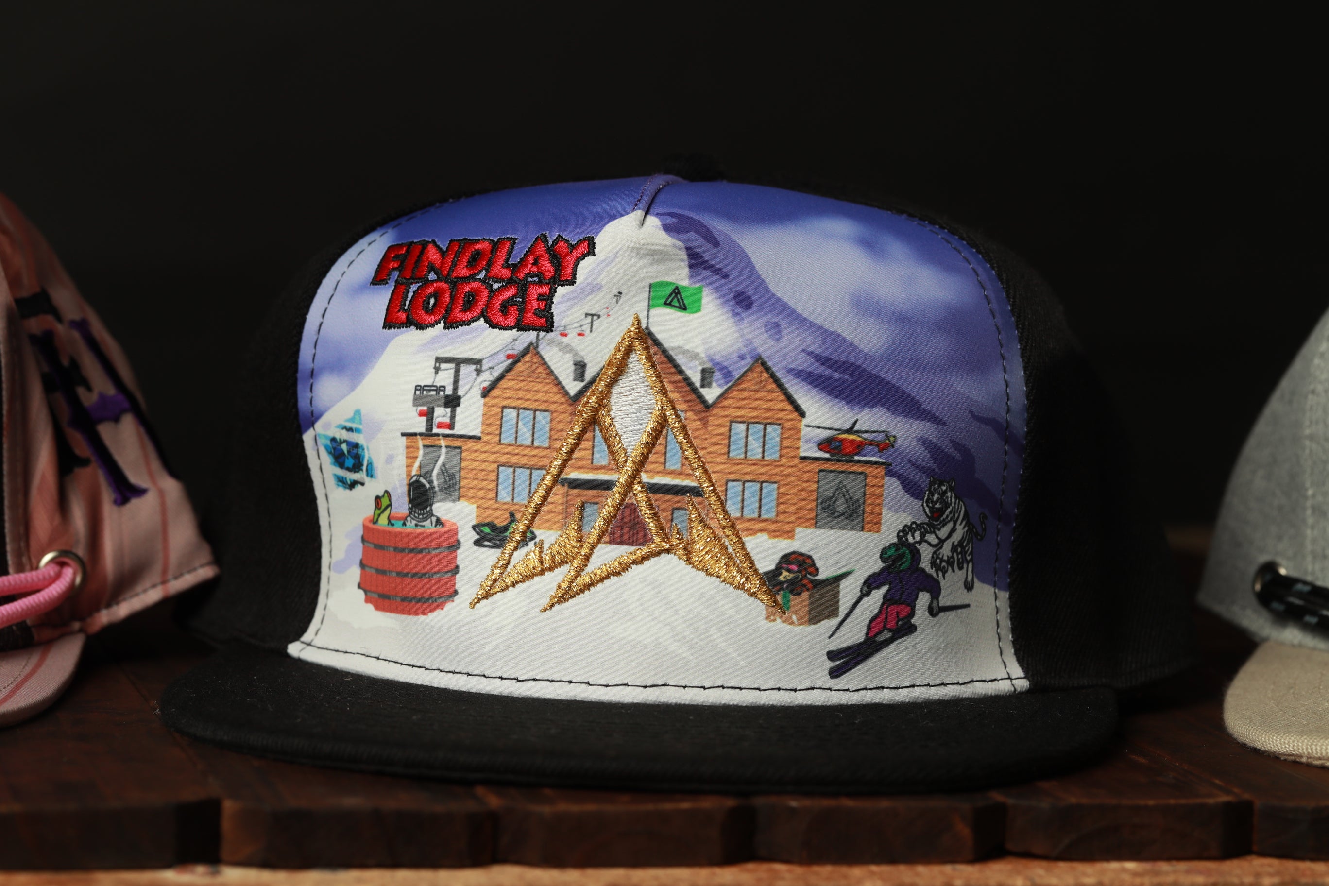 A black baseball cap with a colorful graphic depicting a lodge and ski equipment, along with the text "FINDLAY LODGE".