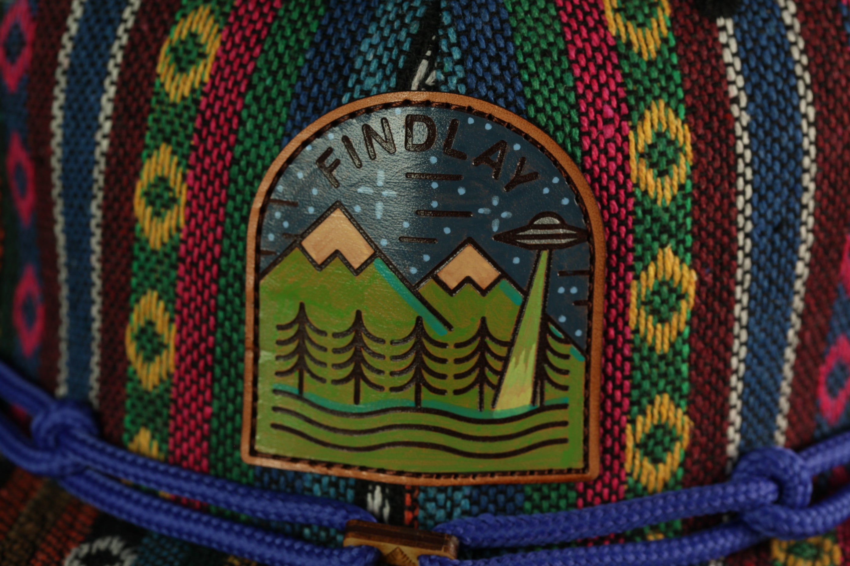 a colorful, patterned fabric with a distinctive embroidered design on it. The design includes a mountain landscape with a small alien spacecraft in the sky, surrounded by trees.