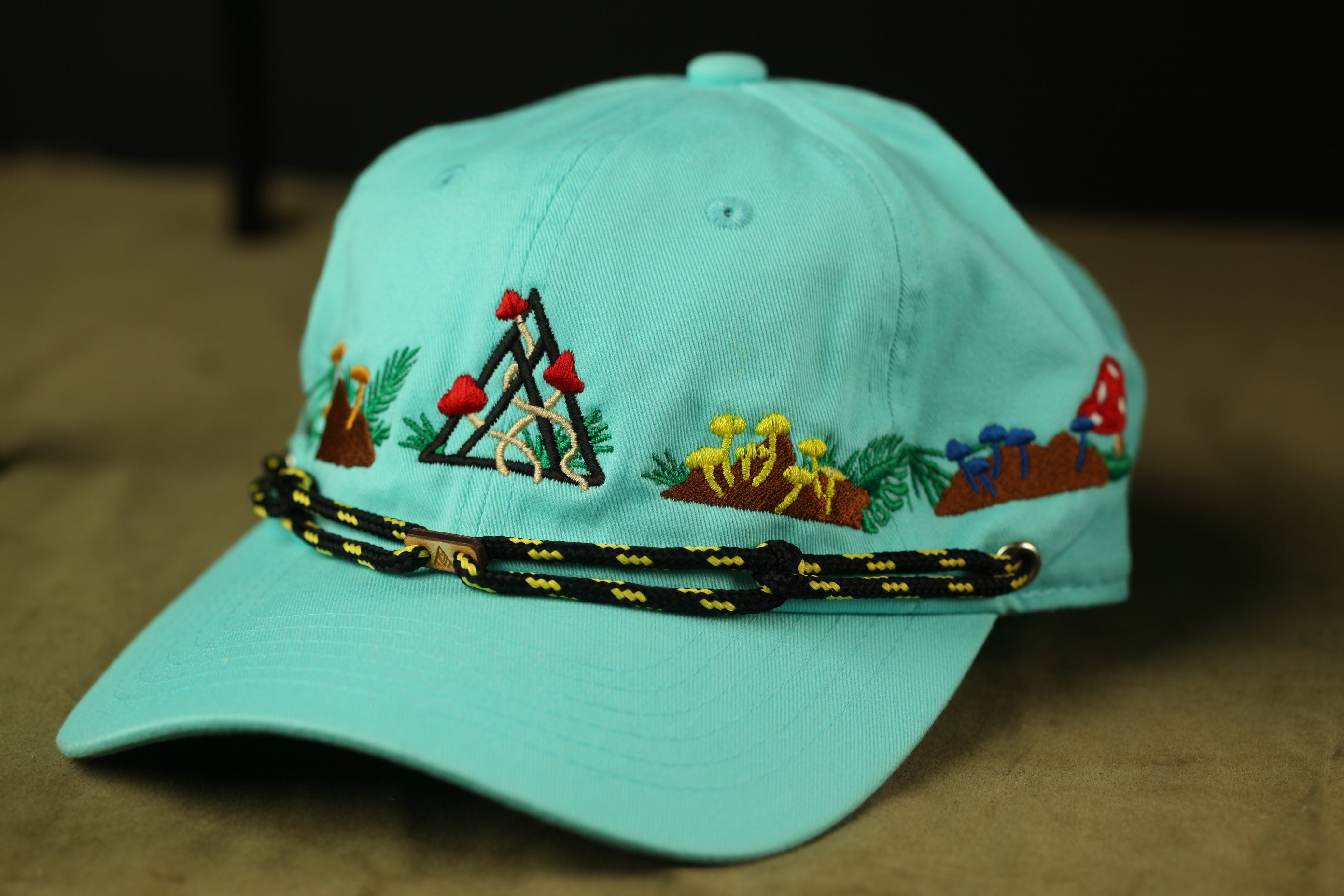 A turquoise baseball cap with a black and yellow rope strap and various colorful mushroom designs on the brim.