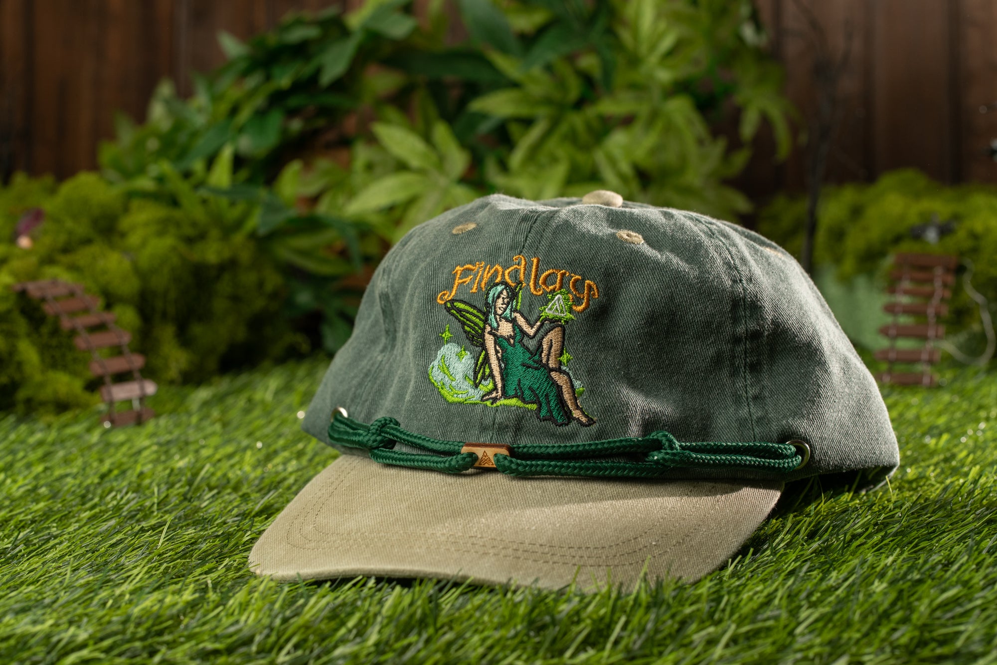 A green baseball cap with a fairy-like design sits on a grassy surface, with a wooden fence and greenery in the background.