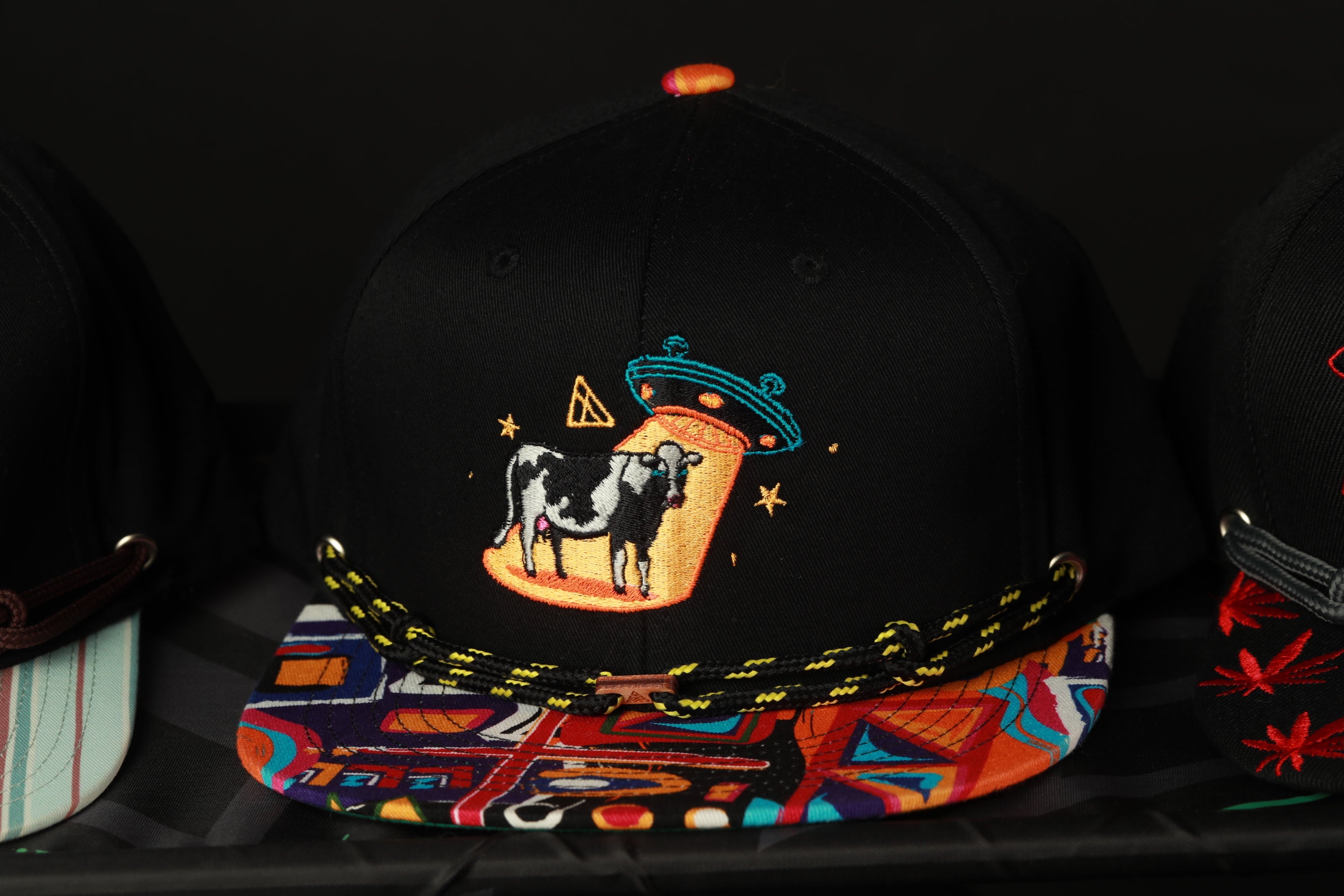 A black baseball cap with a colorful, embroidered design featuring a cow and an alien spacecraft.