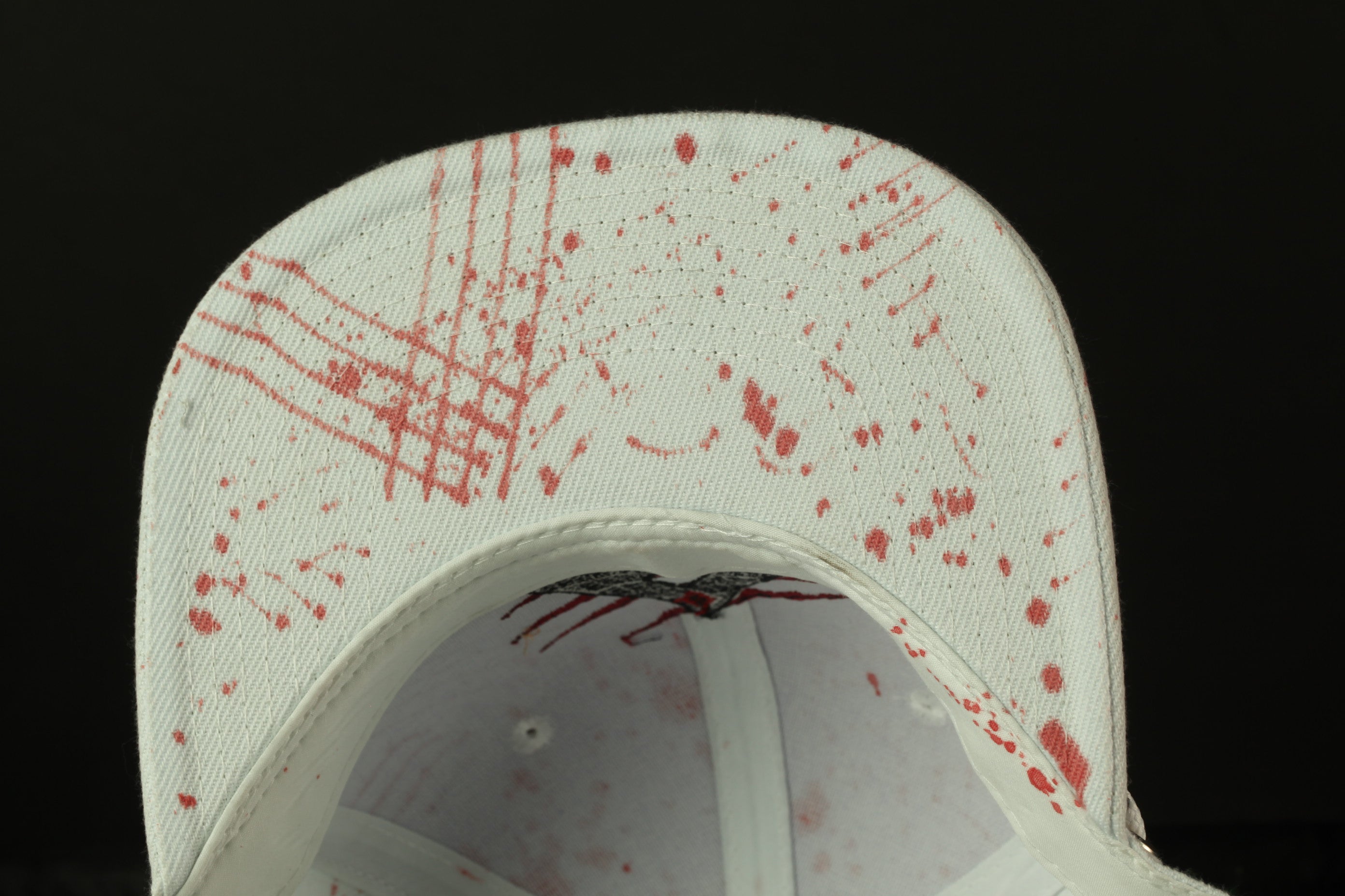 a close-up view of a white baseball cap with red splatters on it, against a dark background.