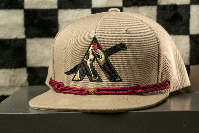 A beige baseball cap with a black and gold logo on the front, featuring a stylized letter 'A' with a knife-like design.
