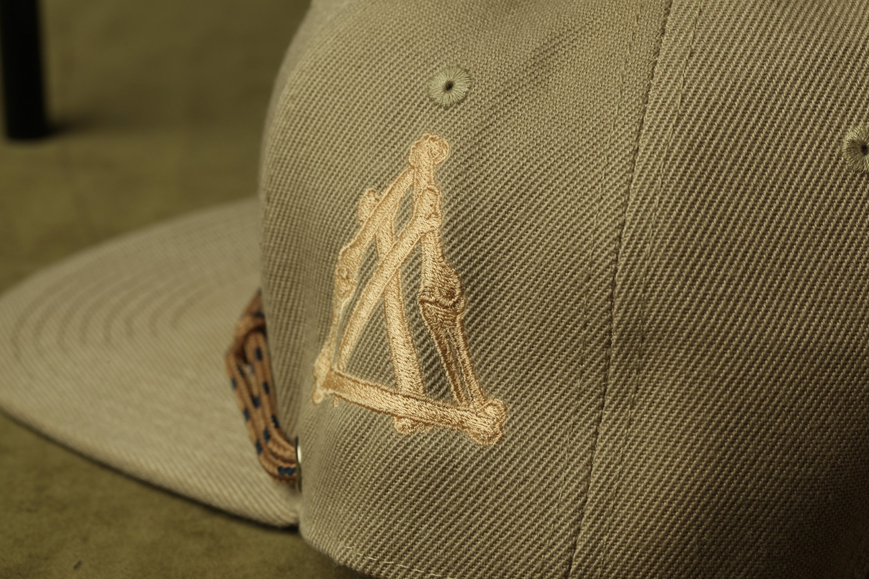 A close-up view of a beige baseball cap with a gold-colored embroidered logo on the front.