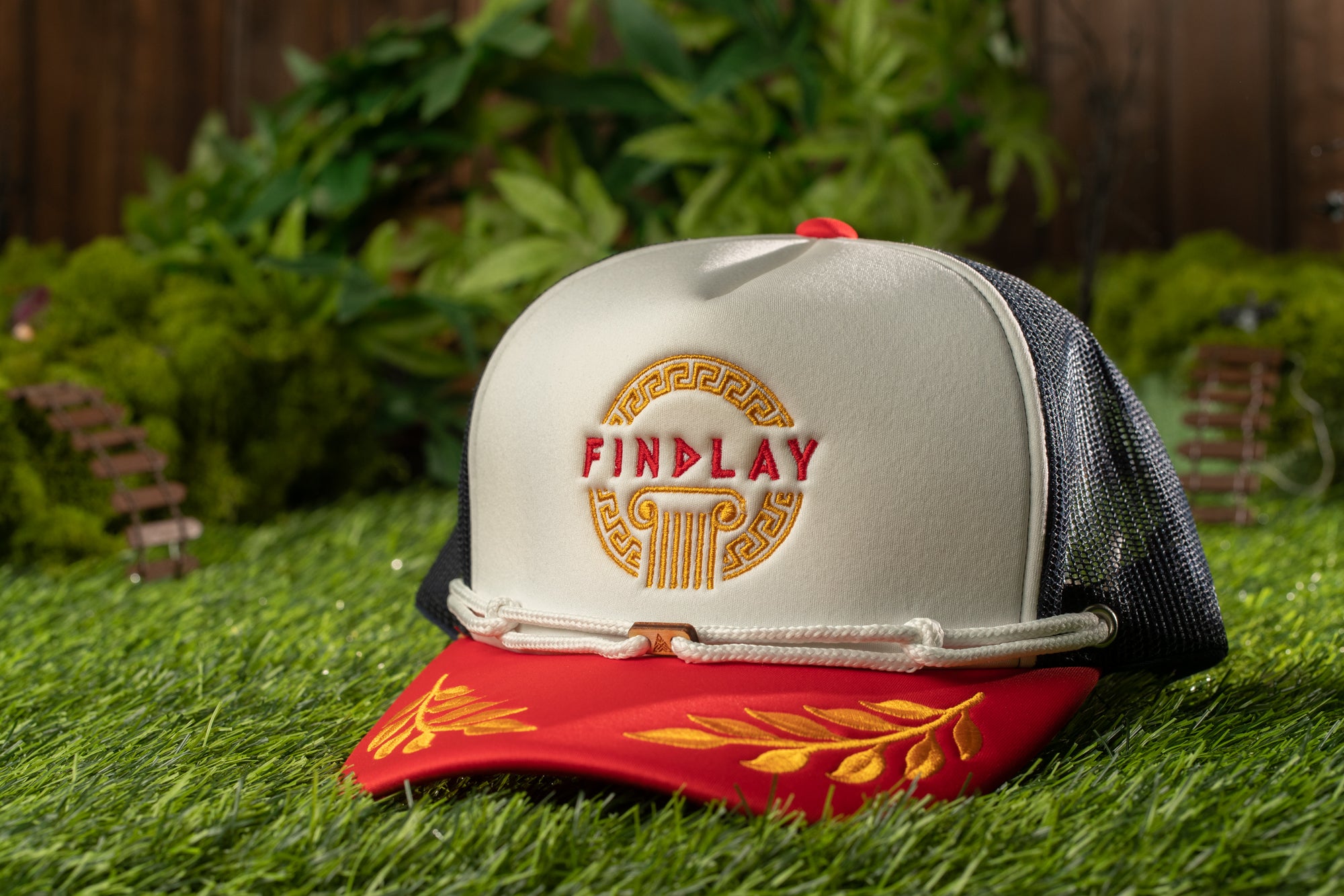 A white baseball cap with a red brim and gold leaf design, featuring the word "FINPLAY" in red letters on the front, is placed on a grassy surface.