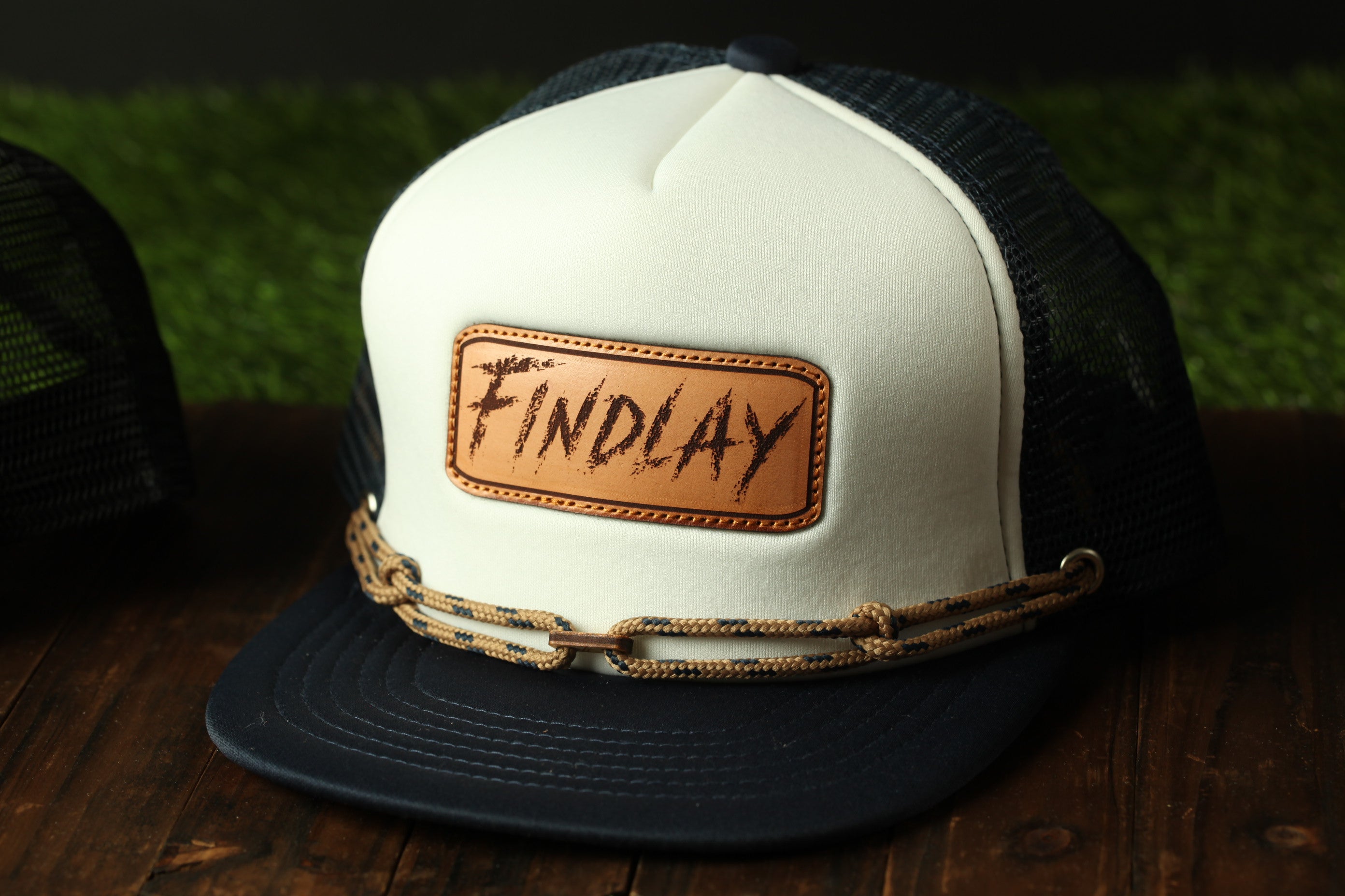 A white baseball cap with a leather patch that says "FINDLAY" on it, placed on a wooden surface.