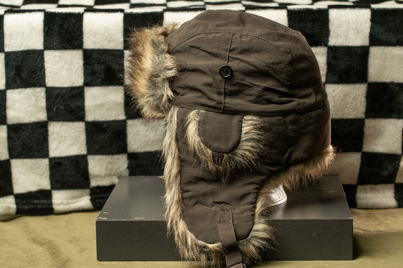 A brown fur-lined winter hat is displayed on a gray surface, with a black and white checkered background.