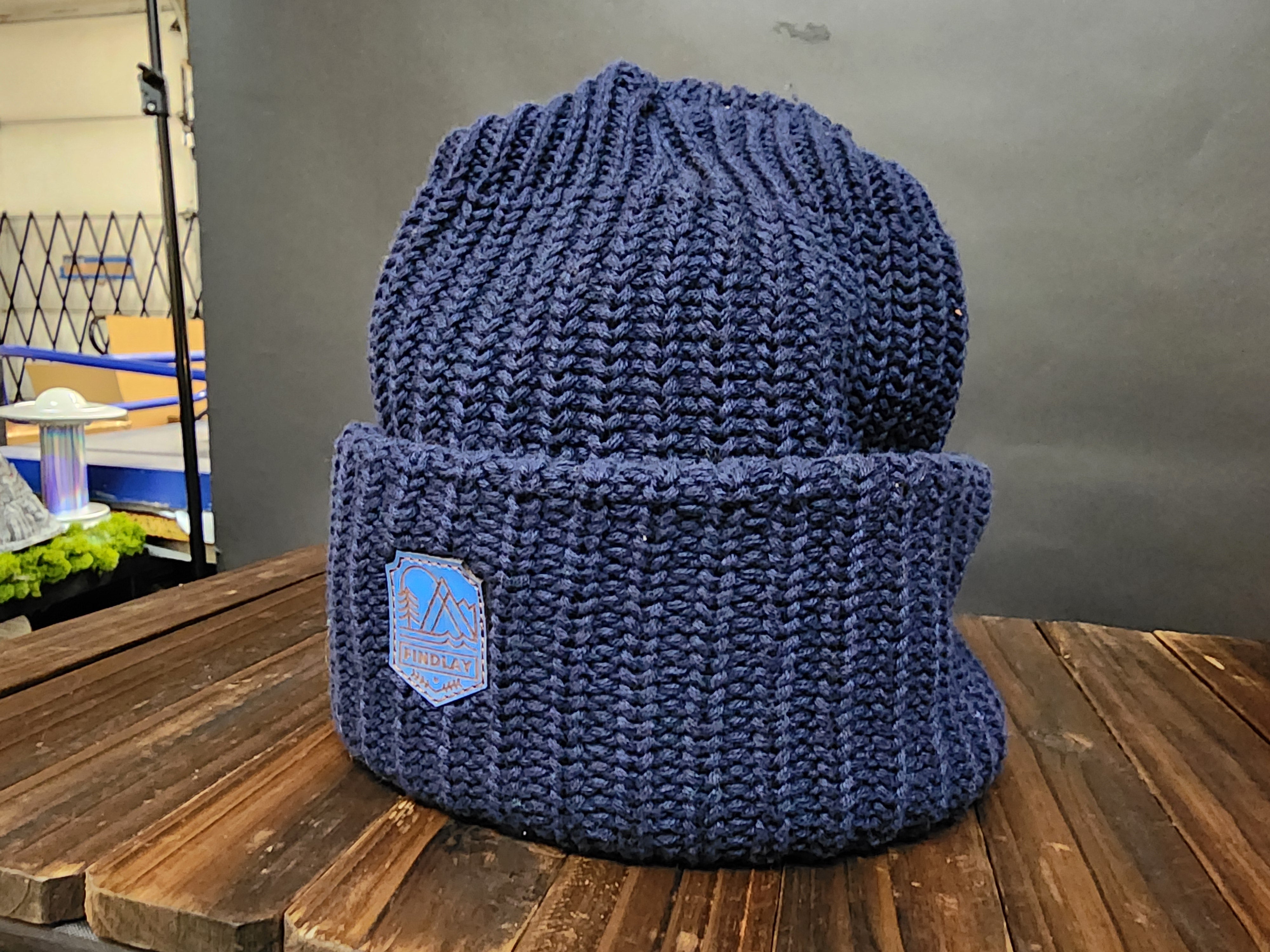 A blue knit beanie with a blue patch on the front, sitting on a wooden surface.