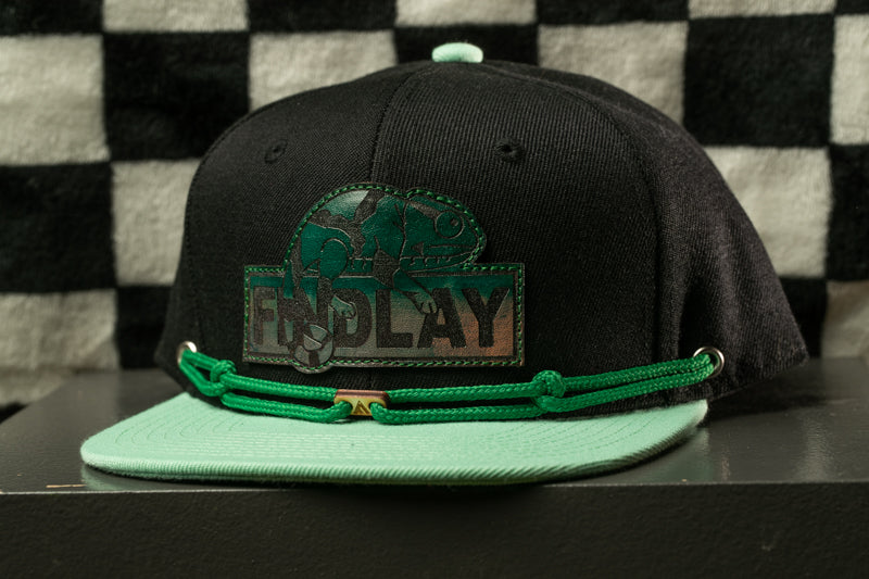 A black baseball cap with a green band and a logo on the front, placed on a black surface against a checkered background.