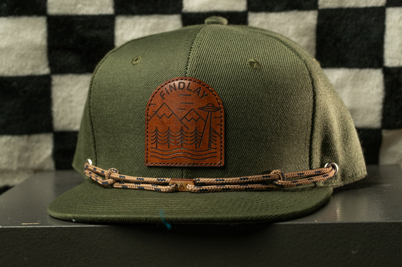 A green baseball cap with a brown leather patch on the front, featuring a mountain landscape design.