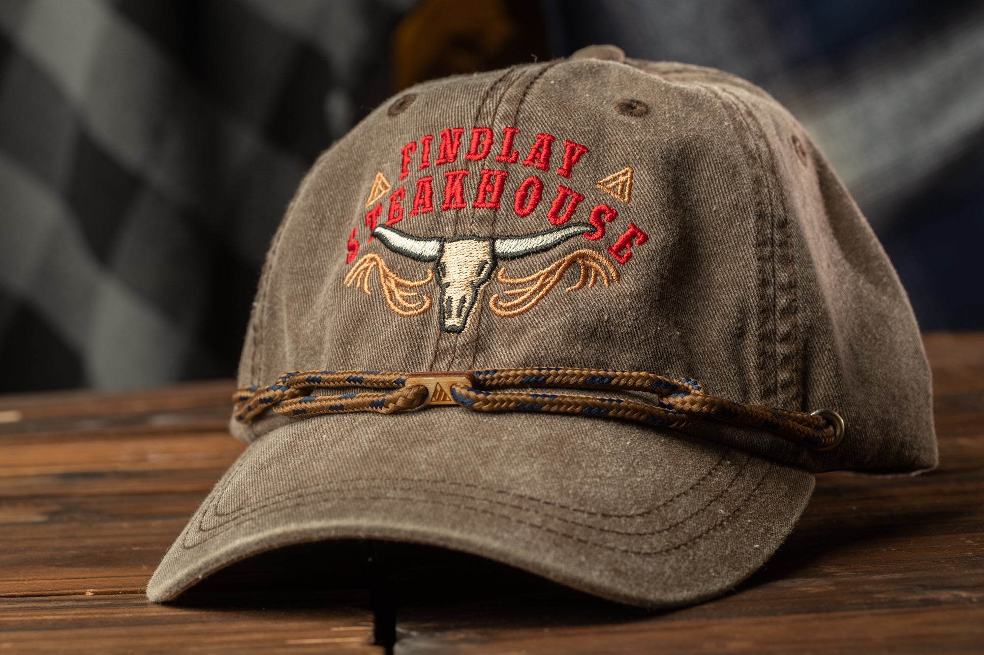 A brown baseball cap with a cowboy logo and the text "FIND LAY STEAKHOUSE" printed on it, sitting on a wooden surface.