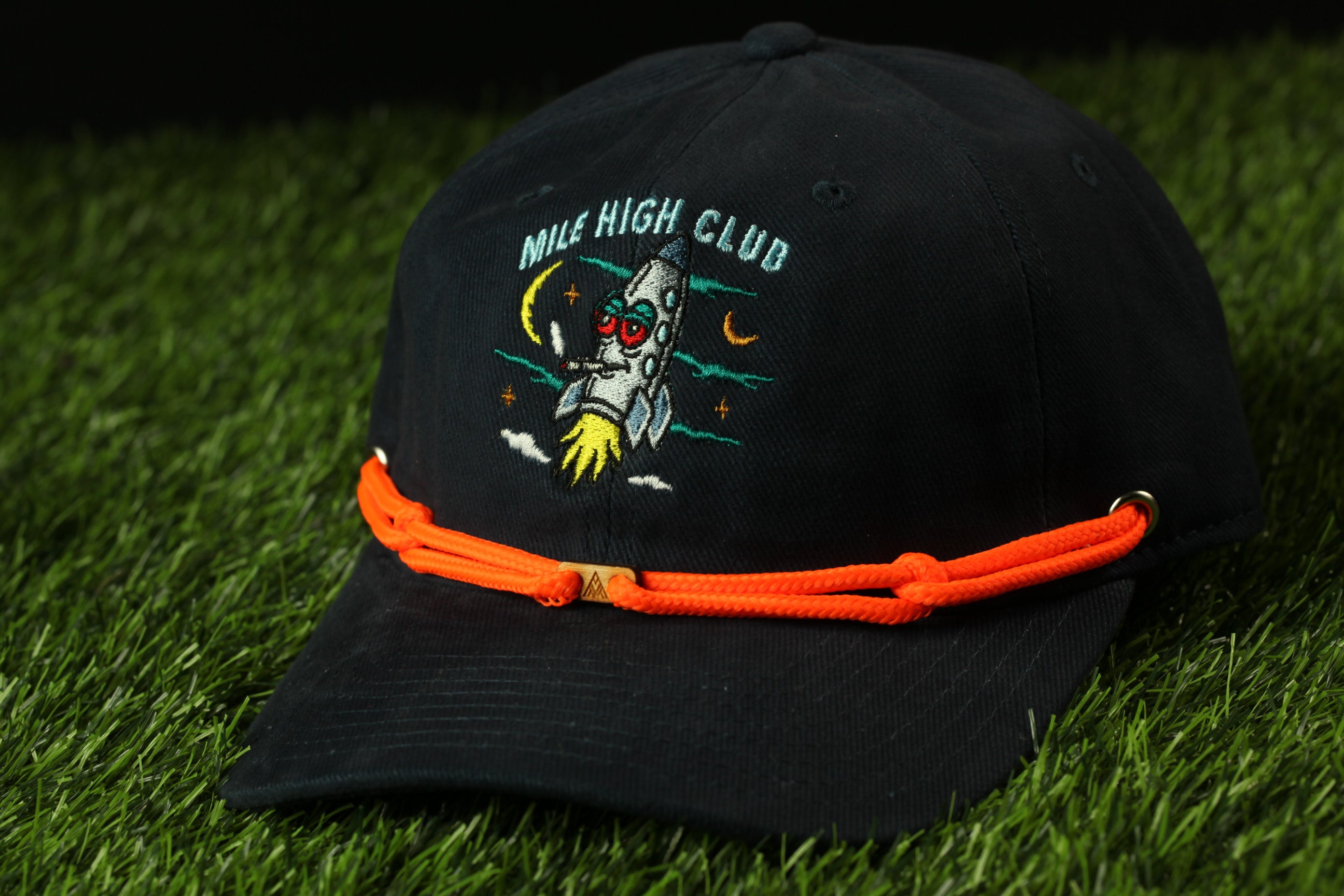 A black baseball cap with an orange string and a rocket ship design on it, placed on a grassy surface.