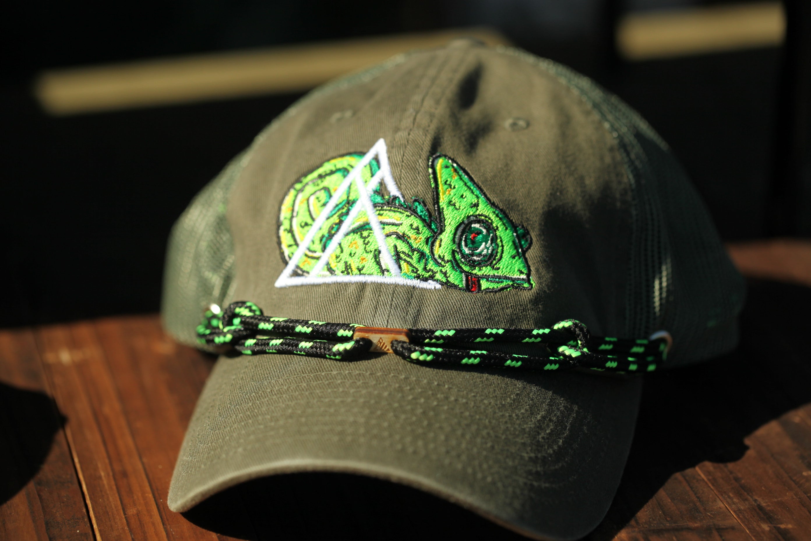 A green baseball cap with a chameleon design on the front, sitting on a wooden surface.