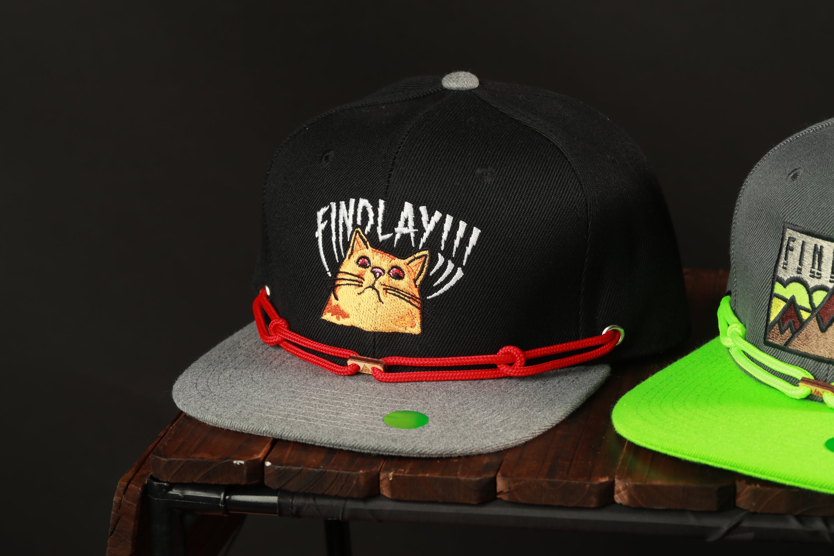 A black baseball cap with a cat design and the text "FIND LAY" printed on it, along with a red string or strap.