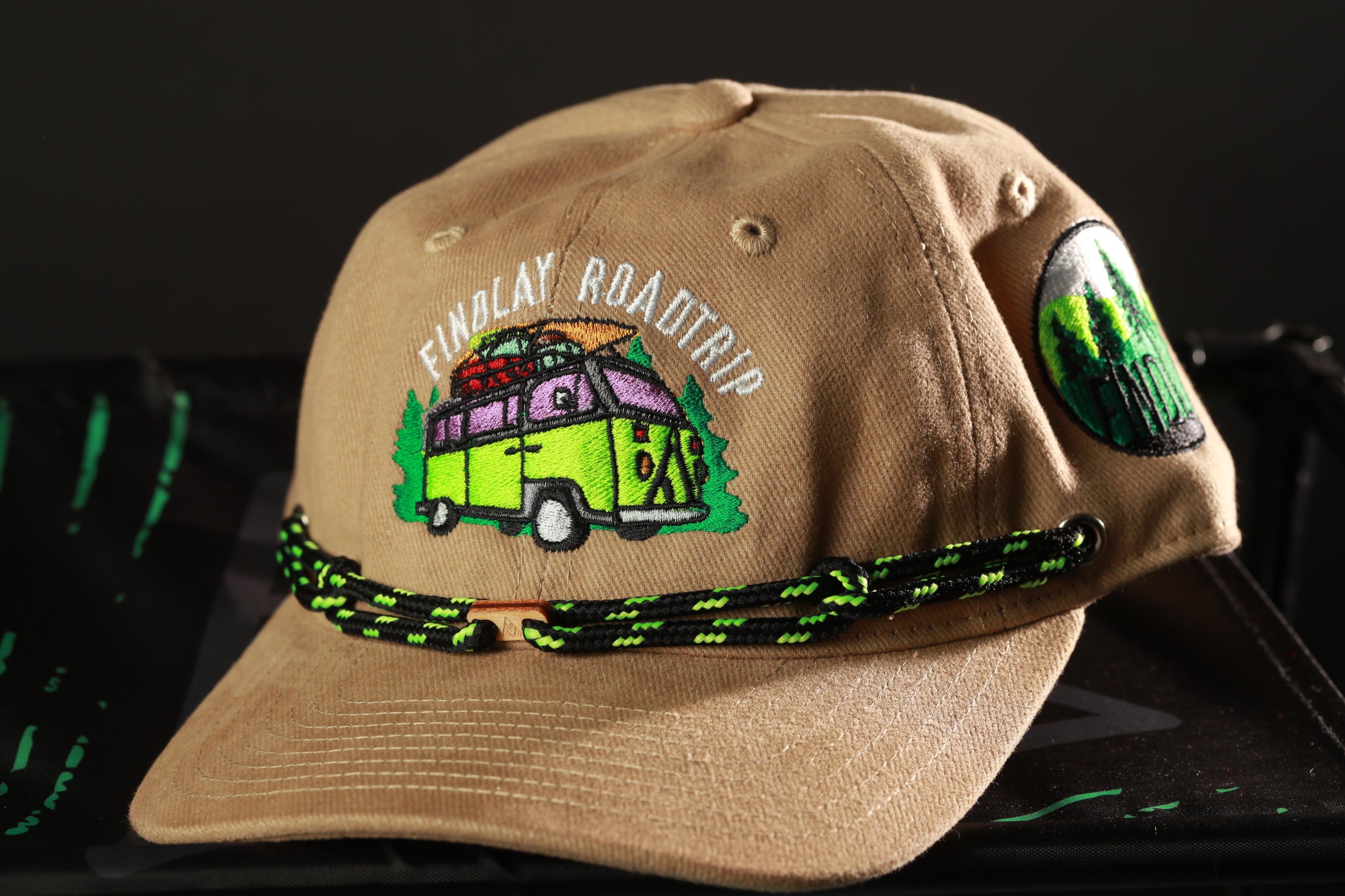 A tan baseball cap with a colorful embroidered design and a green and black string around the brim.