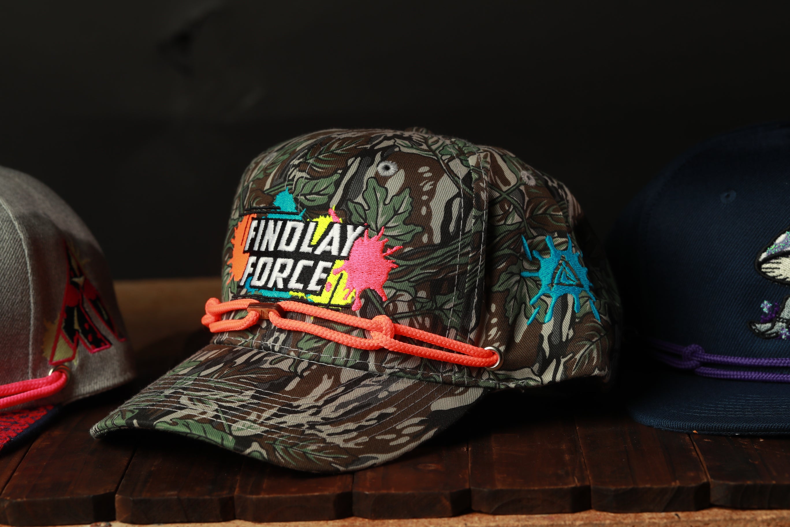 A camouflage baseball cap with the text "FINDAY FORCE" printed on it, along with colorful splatter designs, is displayed on a wooden surface.