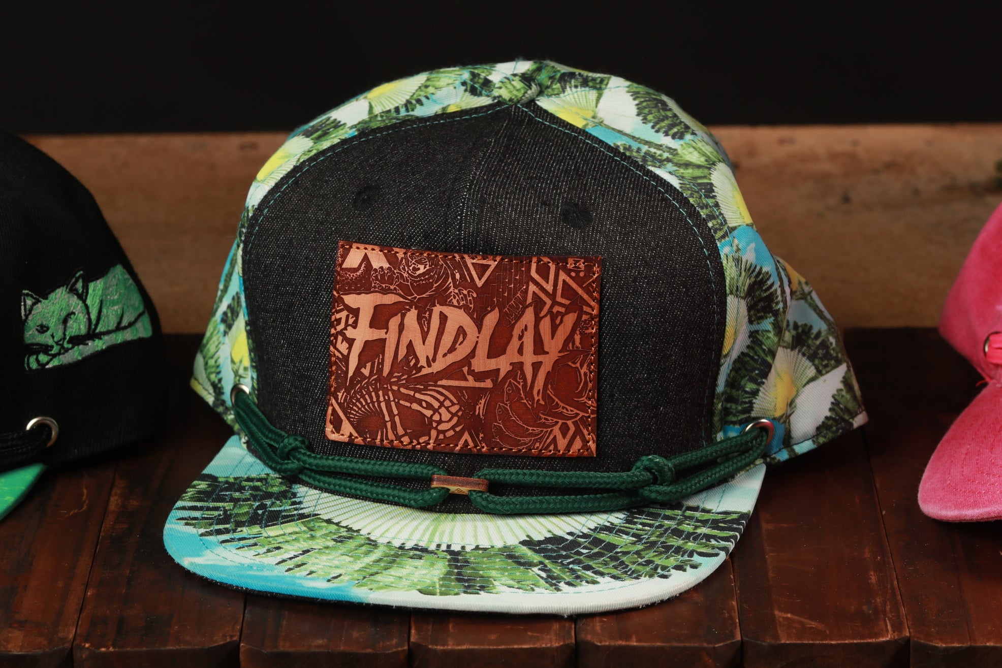 A colorful baseball cap with a tropical print pattern and a leather patch with the word "FINDLAY" on it, displayed on a wooden surface.