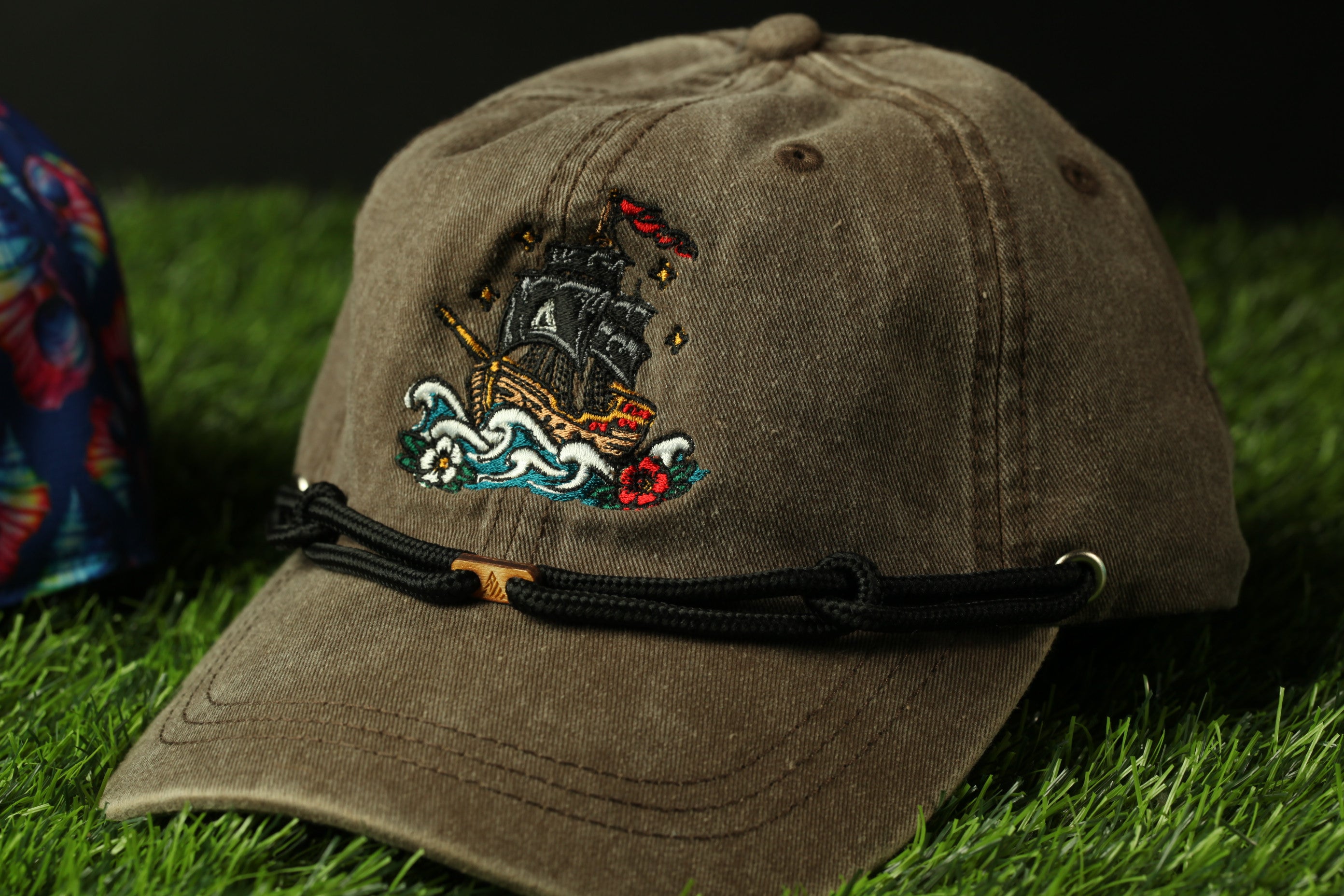 A brown baseball cap with a colorful embroidered design of a ship on the front, resting on a grassy surface.