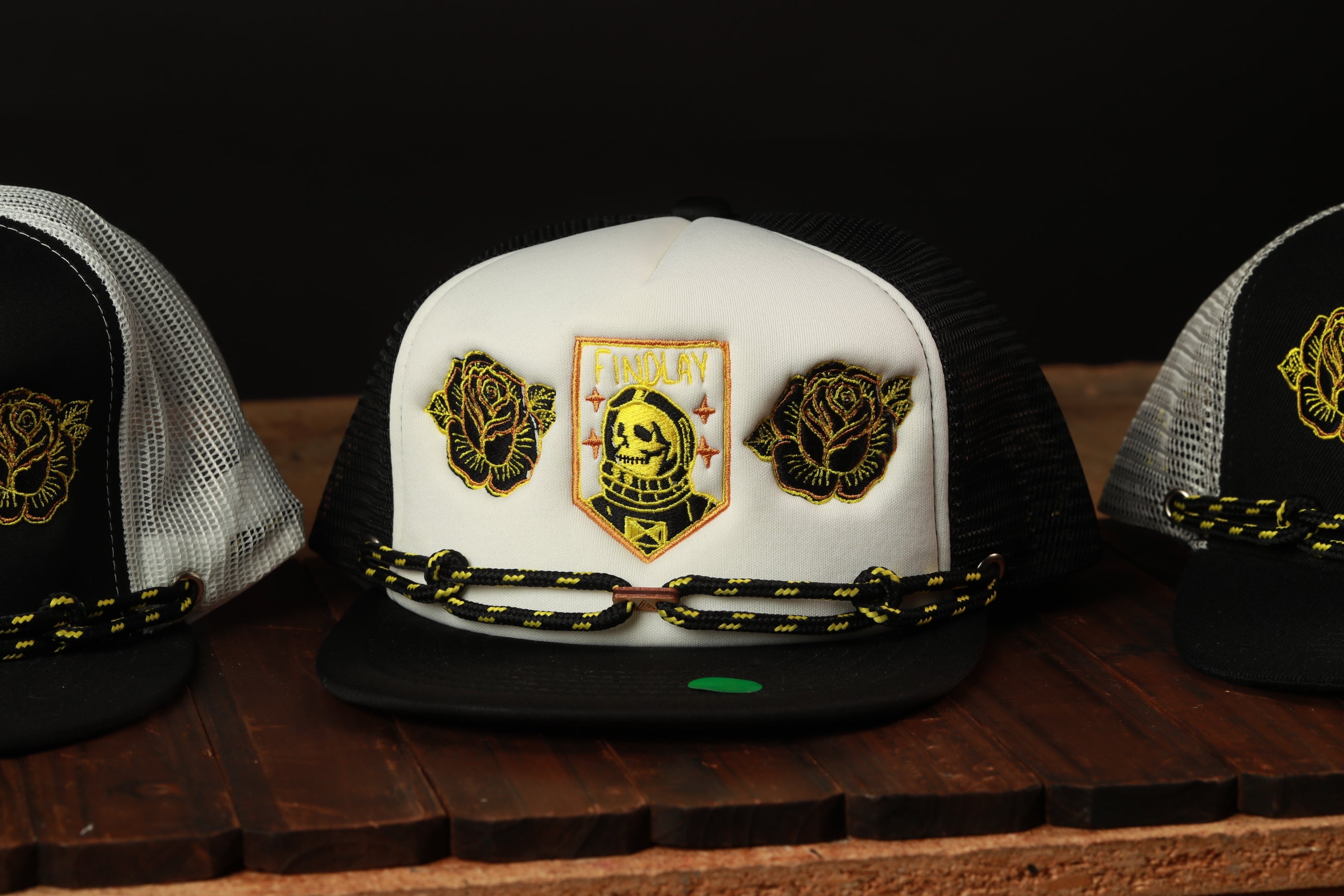 A white baseball cap with black and yellow accents, including a logo and two flower-like patches, is displayed on a wooden surface.